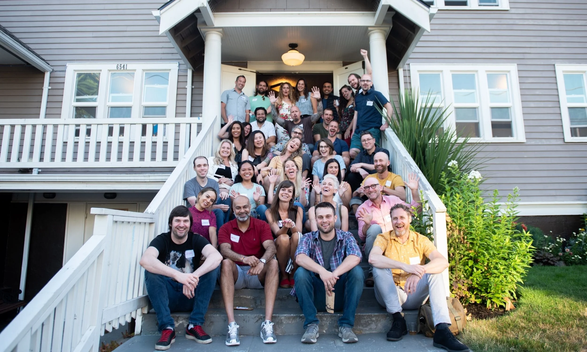 A large group of people sitting and standing on the front steps of a house, smiling and celebrating outdoors.