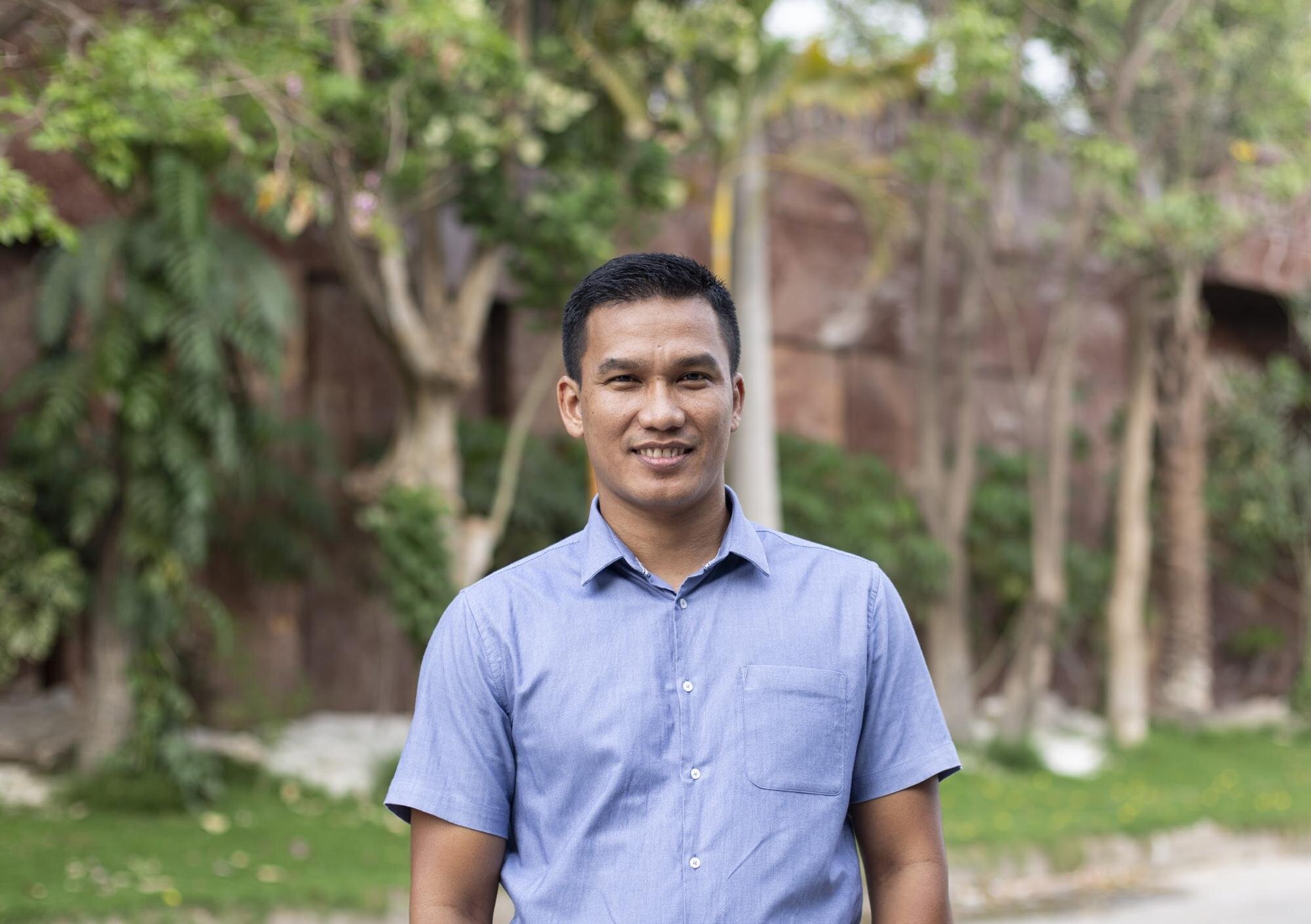 Pannha En, smiling outdoors, standing before trees and greenery, wearing a light-blue button-up shirt.