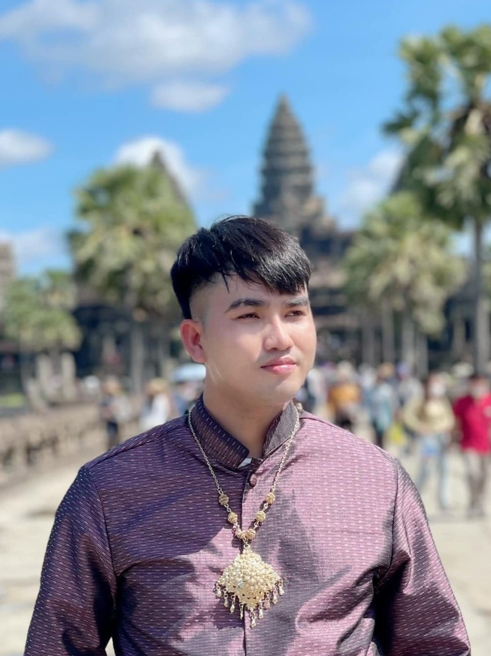 A young man wearing a traditional outfit and jewelry standing outdoors near a historic temple, with trees and a blue sky in the background.