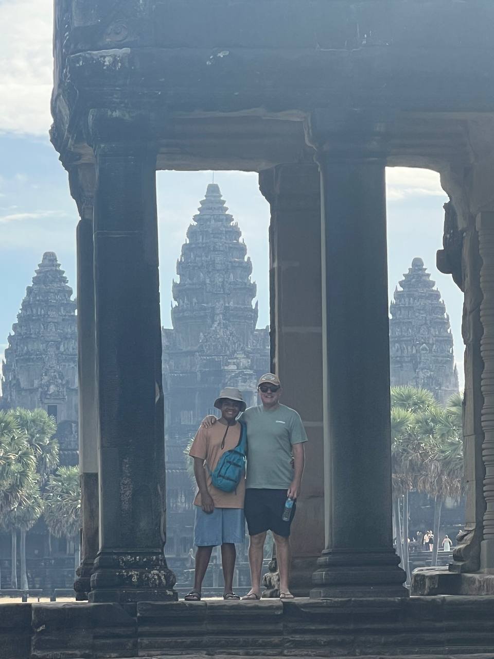 Two tourists standing under an ancient stone archway with temple towers in the background, likely at Angkor Wat in Cambodia.