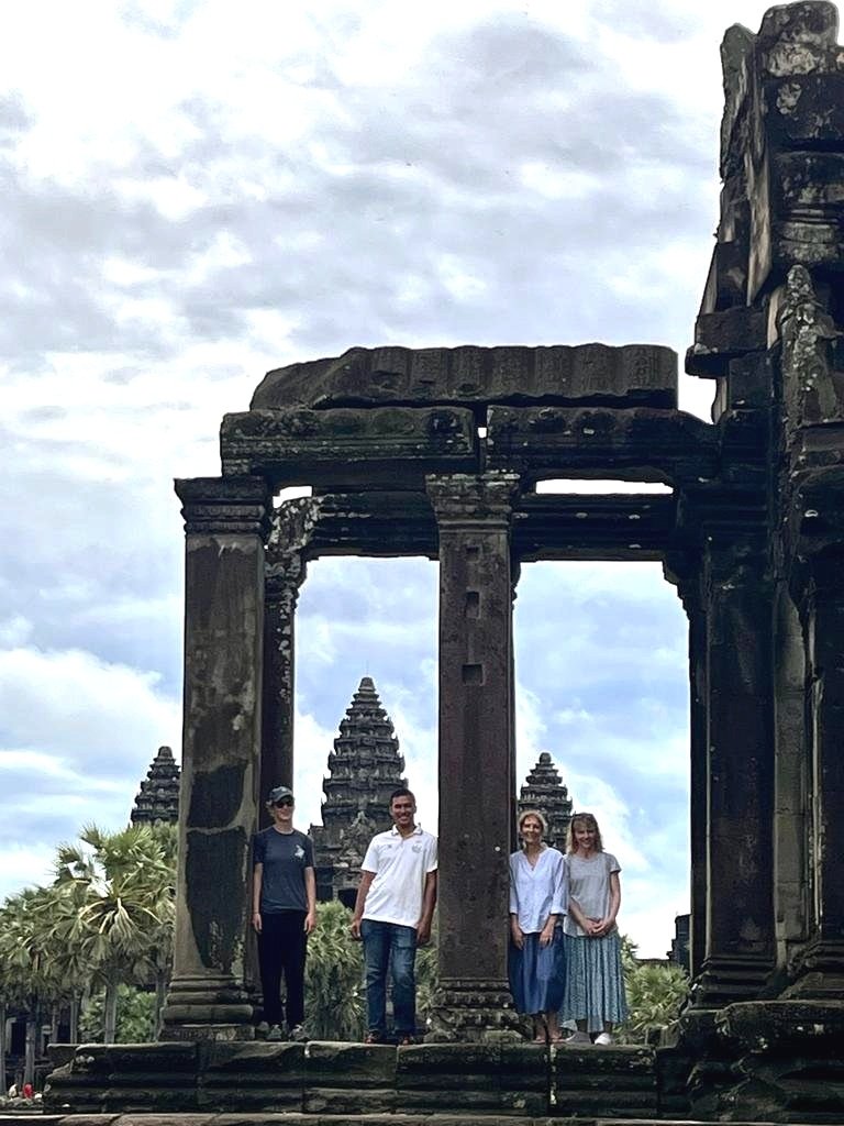 Four people standing under a stone archway in front of ancient temple ruins with palm trees in the background.