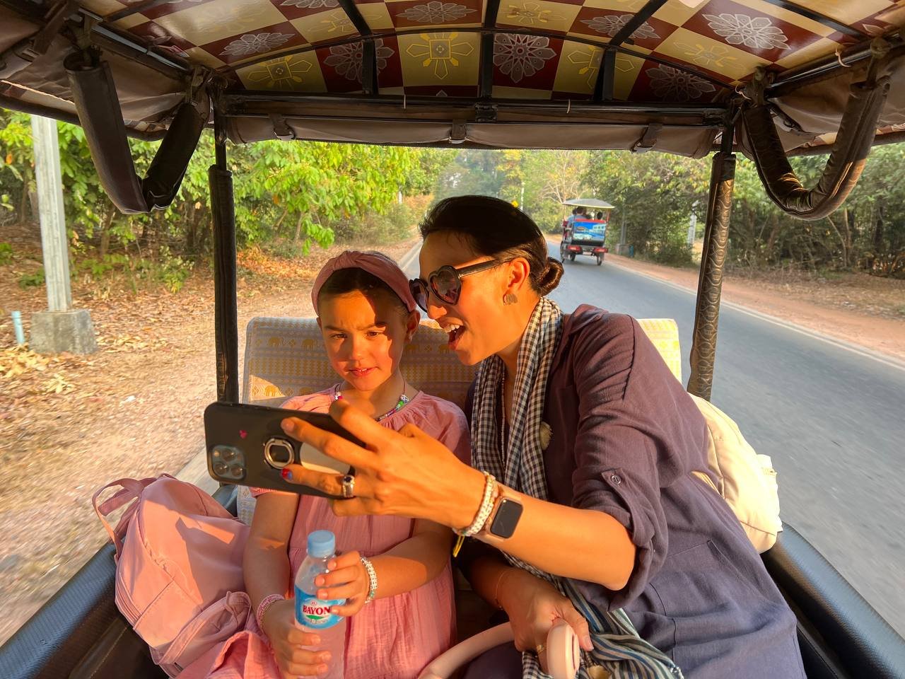 A woman and a young girl are sitting in a tuk-tuk on a road with trees on both sides. The woman is showing something on her phone to the girl, who is holding a water bottle and looking confused. The woman is wearing sunglasses, a gray jacket, and jewelry. The girl is wearing a pink dress and a headband.