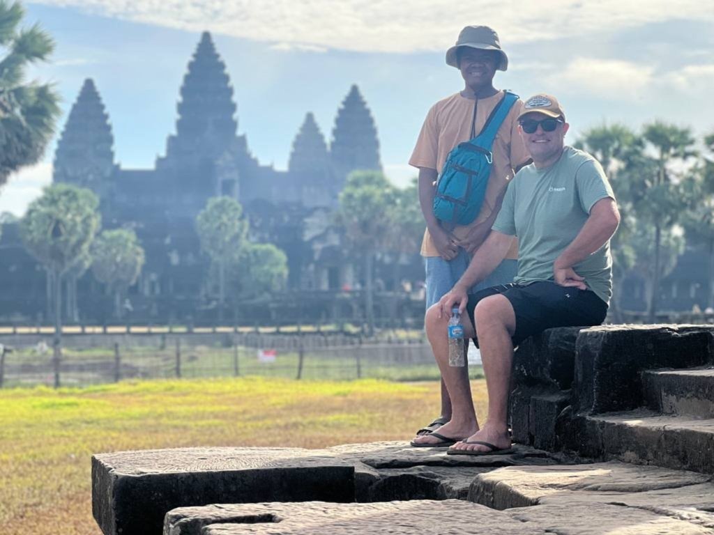 Two men posing on steps in front of Angkor Wat temple in Cambodia on a cloudy day, with trees and the temple visible in the background.