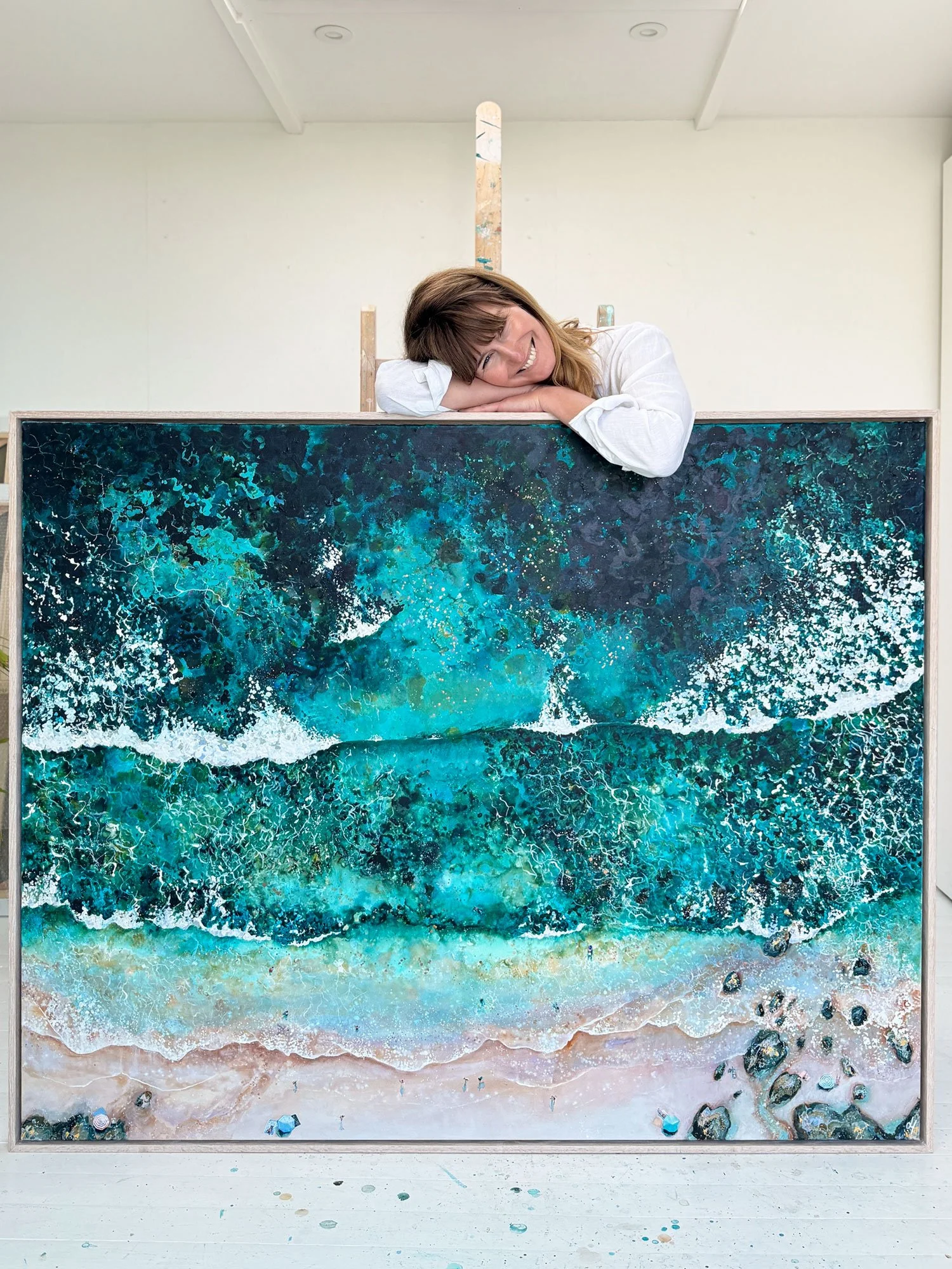British artist Lucy Young in her studio, leaning over a large-scale aerial seascape painting titled 'Breath of Life', featuring dramatic turquoise ocean waves and 24k gold leaf details.