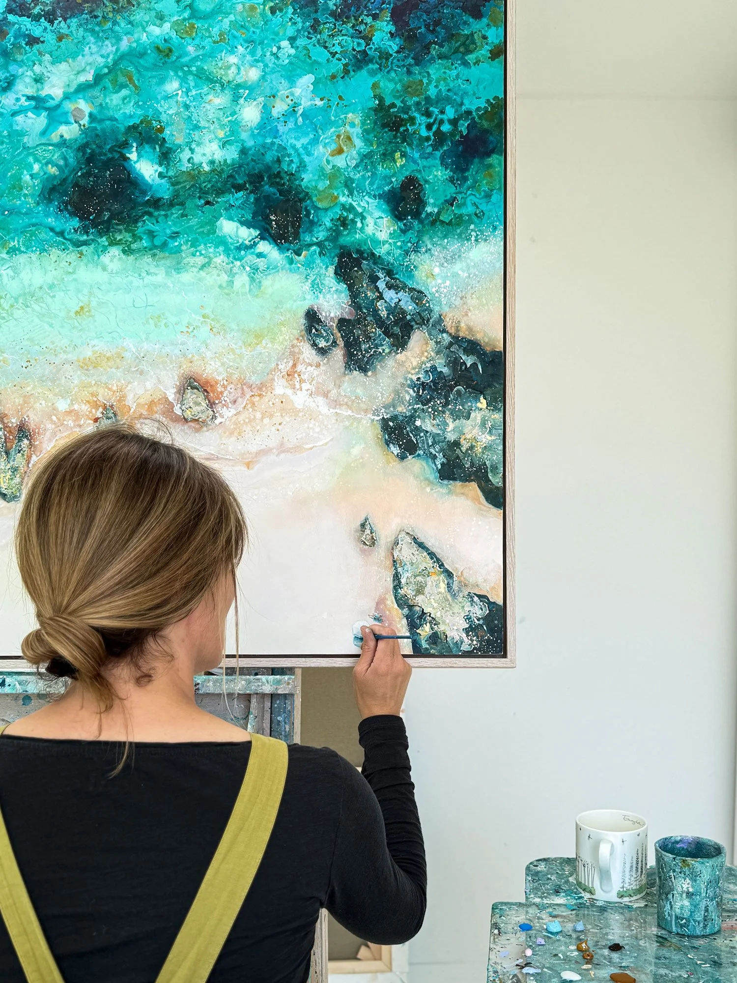 Artist Lucy Young using a fine brush to paint subtle characters into a bespoke aerial seascape commission, showing the detailed process of creating a custom family beach painting.