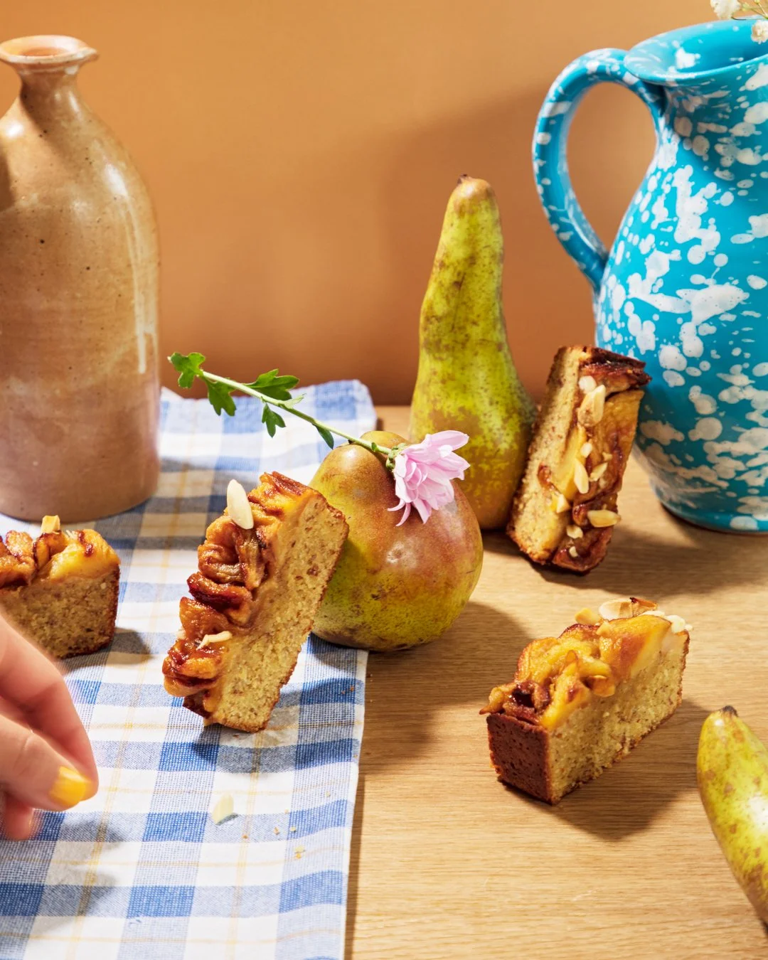 Homemade apple cake slices on a checkered cloth, with fresh pears and a blue and white speckled pitcher in the background.
