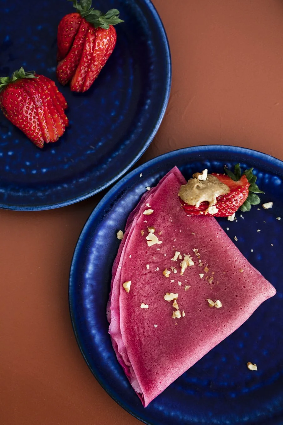 Two blue plates with strawberries; one plate holds three whole strawberries, the other a pink crepe with strawberries, nut butter, and chopped nuts.