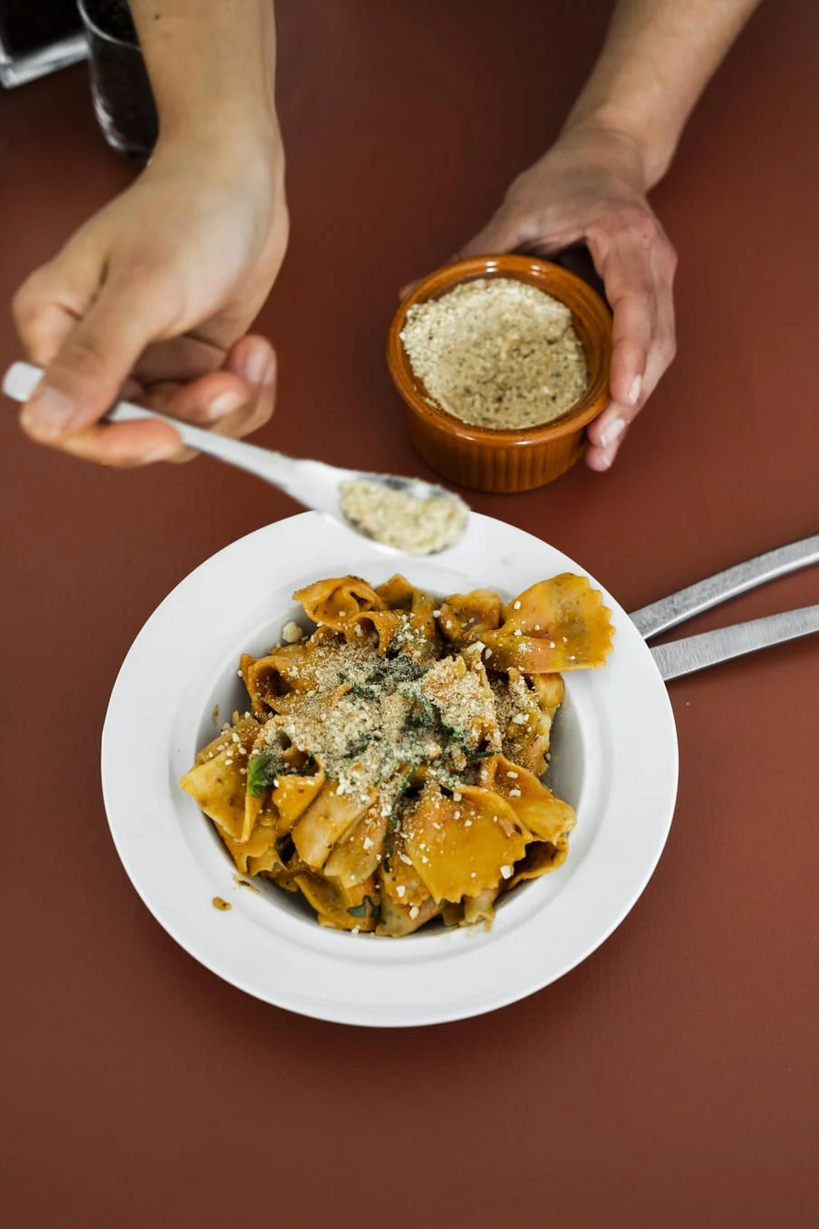 Top-down view of a bowl of cooked pasta with grated cheese and herbs, placed on a red-brown table. A hand is holding a spoonful of grated cheese over the pasta, and another hand is holding a small jar of grated cheese or similar seasoning.