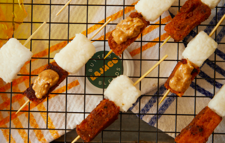 Skewers with fried cubes of fish or chicken topped with peanut sauce, placed on a cooling rack over decorative cloths.