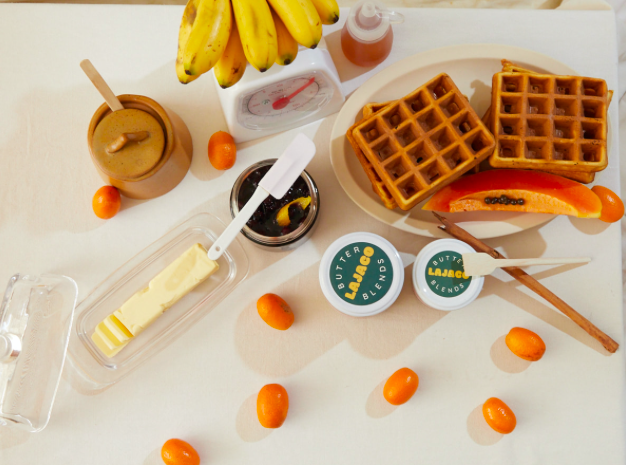 A breakfast scene with bananas, waffles, butter, syrup, and small orange fruits on a white table.