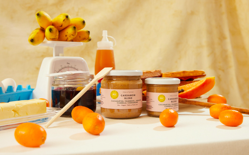 A table with bananas, jars of cardamom spice, papaya, kumquats, honey, and some other food items.