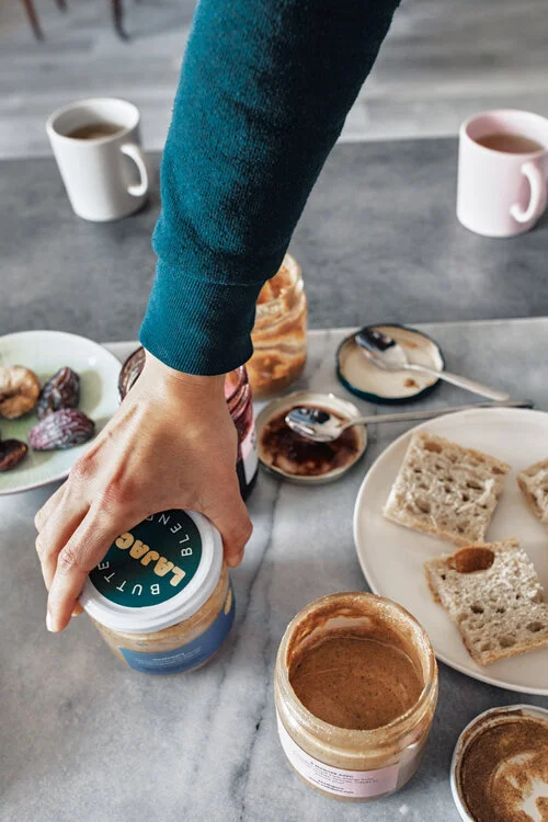 Person spreading peanut butter on bread at breakfast table with coffee, tea, and snacks.