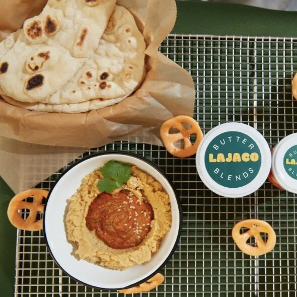 A plate of hummus with a red sauce in the center, garnished with a sprig of cilantro. To the left, a basket lined with parchment paper holds topped flatbread or naan. To the right, there are small containers labeled 'Butter Bajago Blends' and some pr