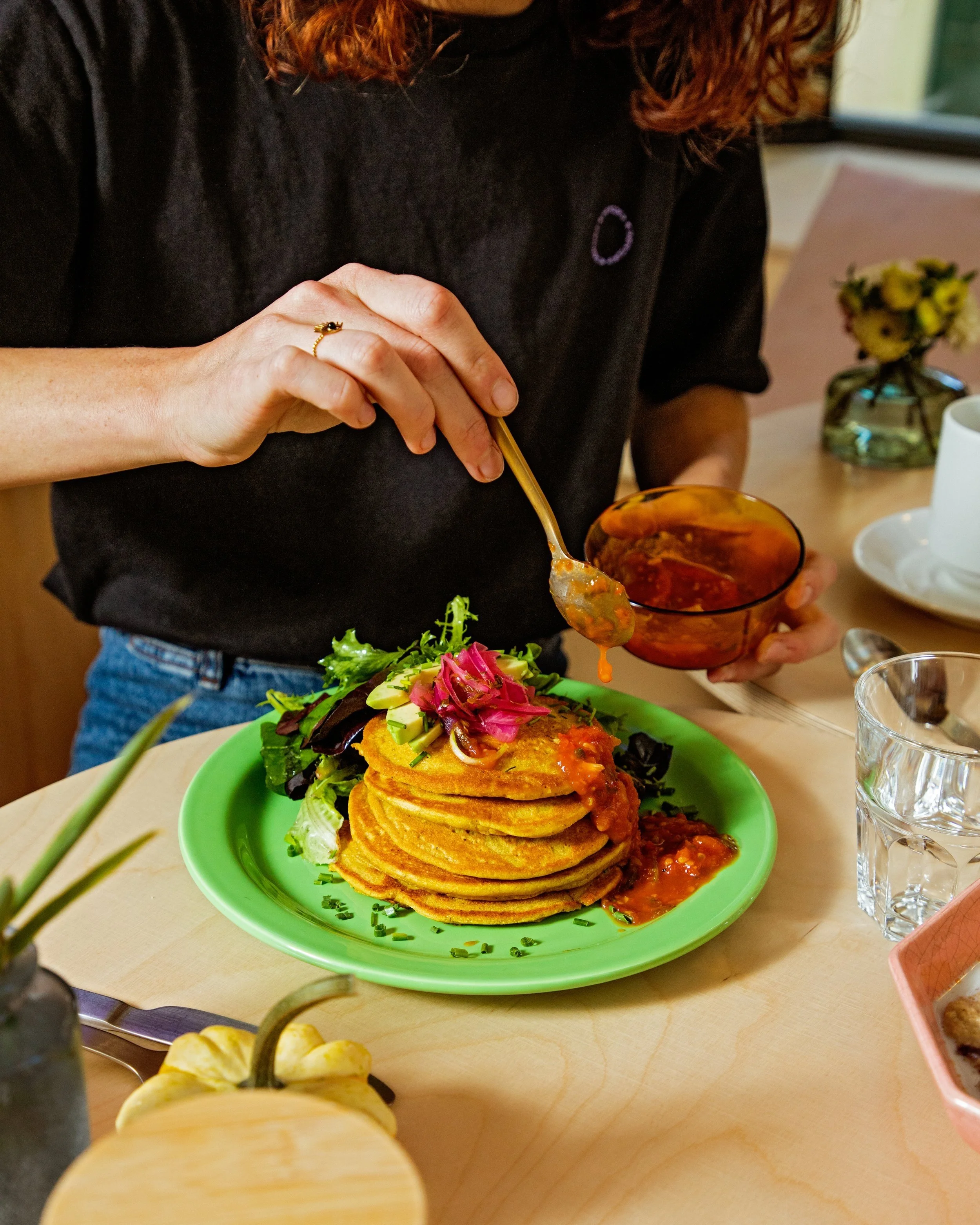 Person with long, red hair wearing a black shirt serving food from a bowl onto a green plate of stacked fried patties topped with fresh vegetables. In the background, there is a small vase with yellow flowers, a cup, and a glass of water on the table