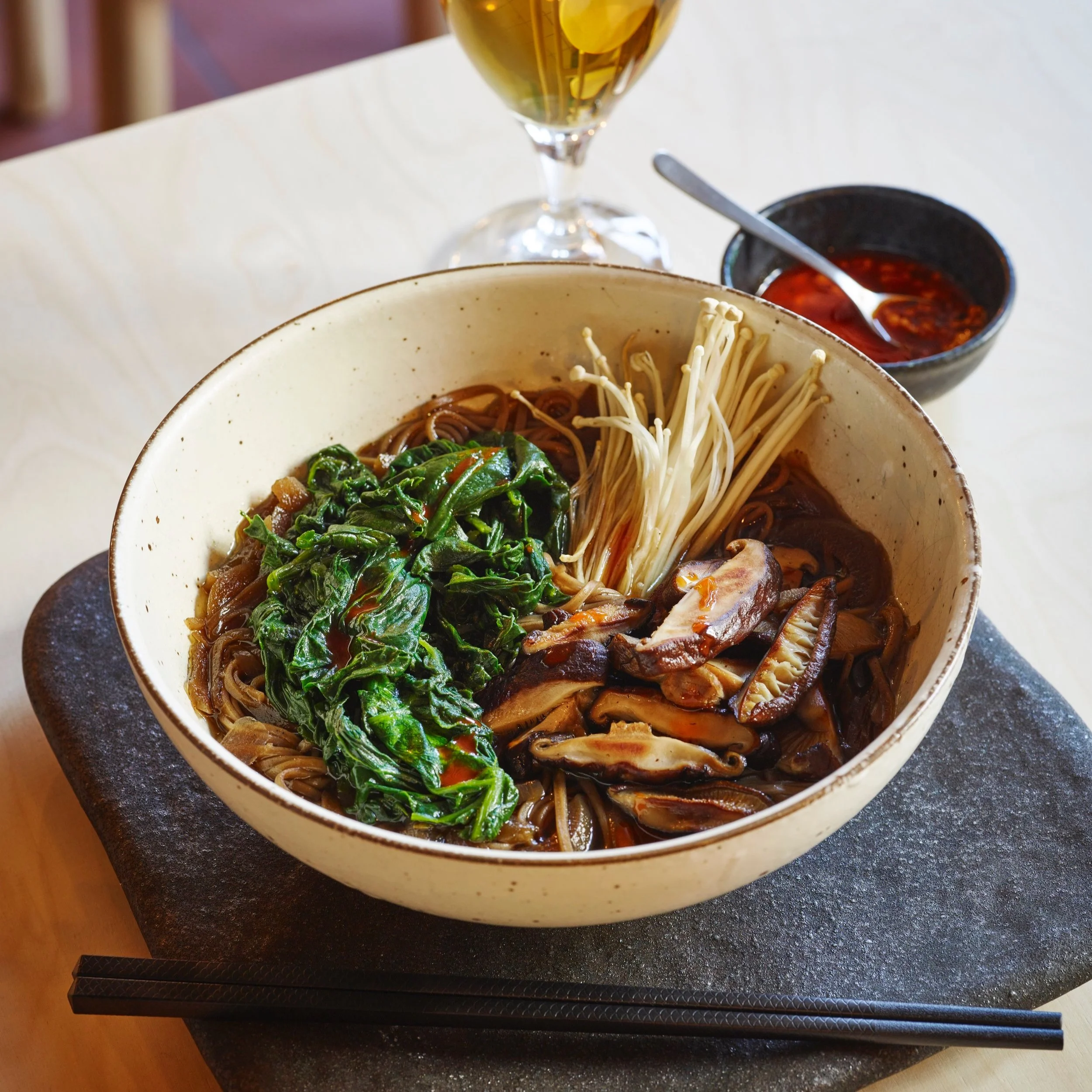 A bowl of Asian noodle soup with spinach, enoki mushrooms, and other vegetables, accompanied by a small bowl of red chili sauce and a glass of white wine with ice cubes on a white table.