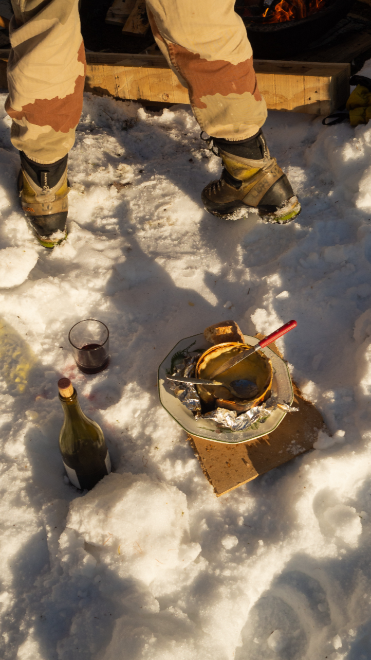 Antique fondue pot melted over a small outdoor fire on snow, with a spoon inside, a piece of bread, a glass of red wine, and a bottle of wine nearby.