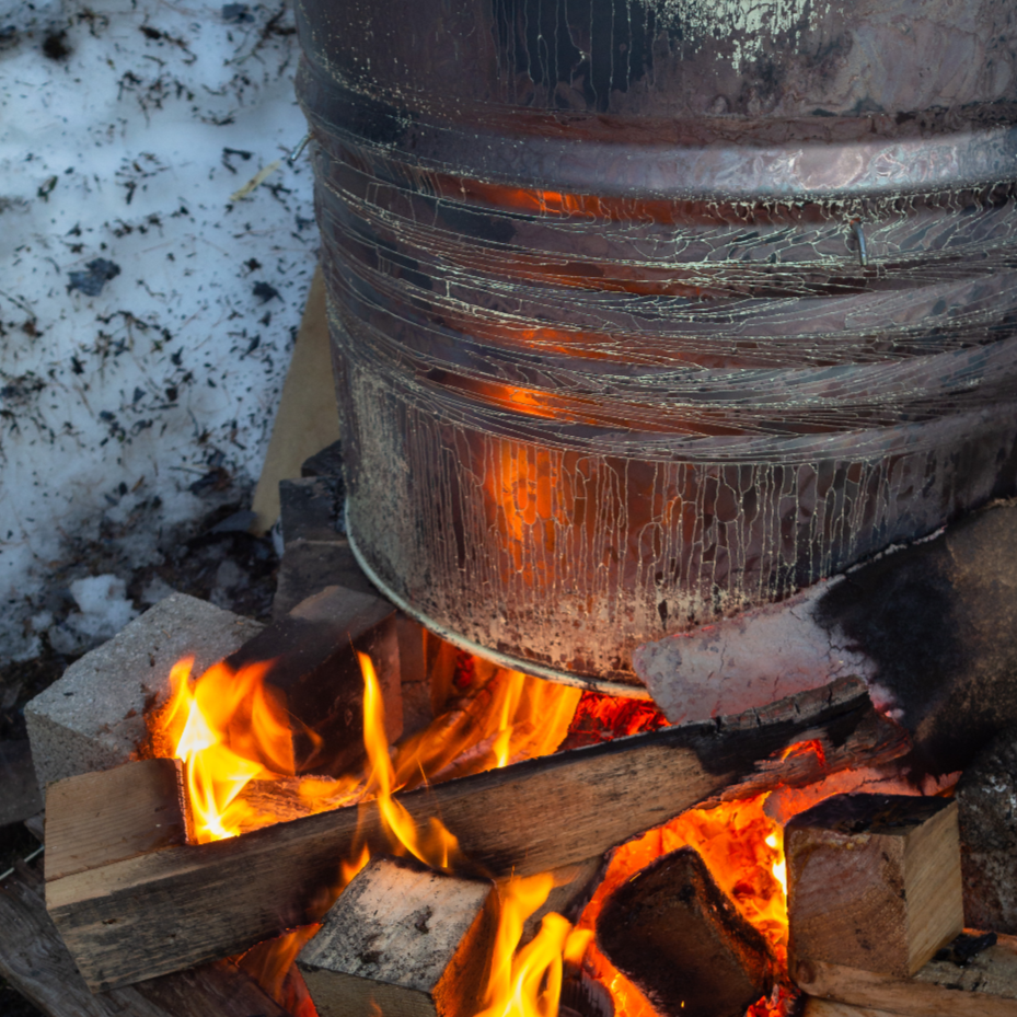 A large metal container placed above an outdoor wood fire, with flames and burning wood below. Snow surrounds the setup.