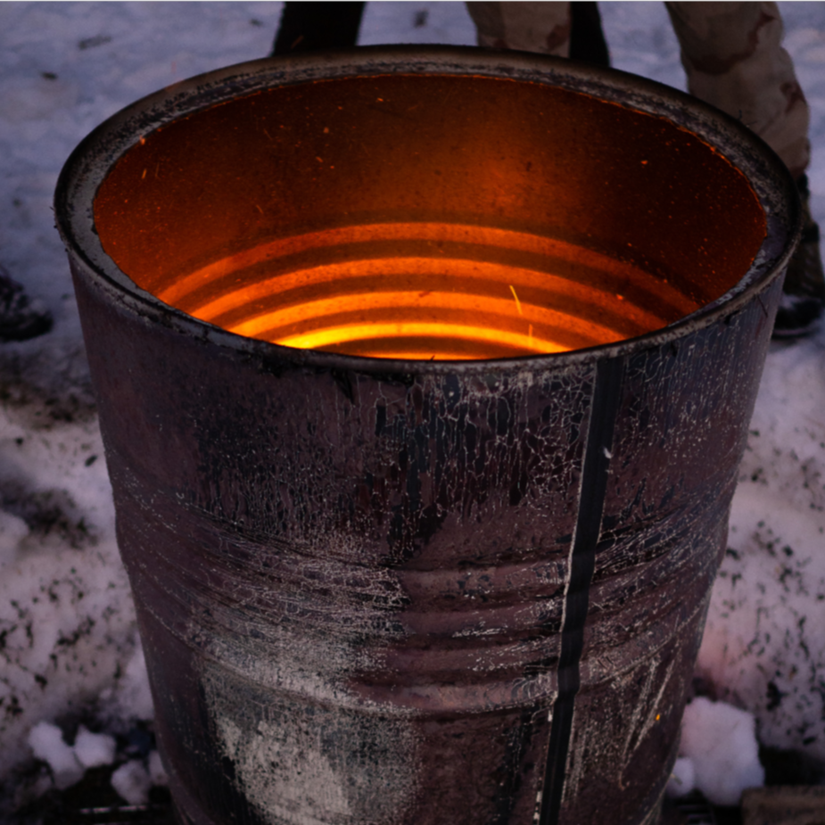 A close-up of a glowing barrel furnace used for heating or melting metal, with snow on the ground around it.