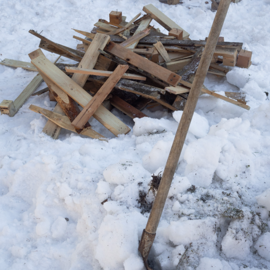 Pile of broken wooden sticks and planks in snow with a long wooden stick leaning against the pile.