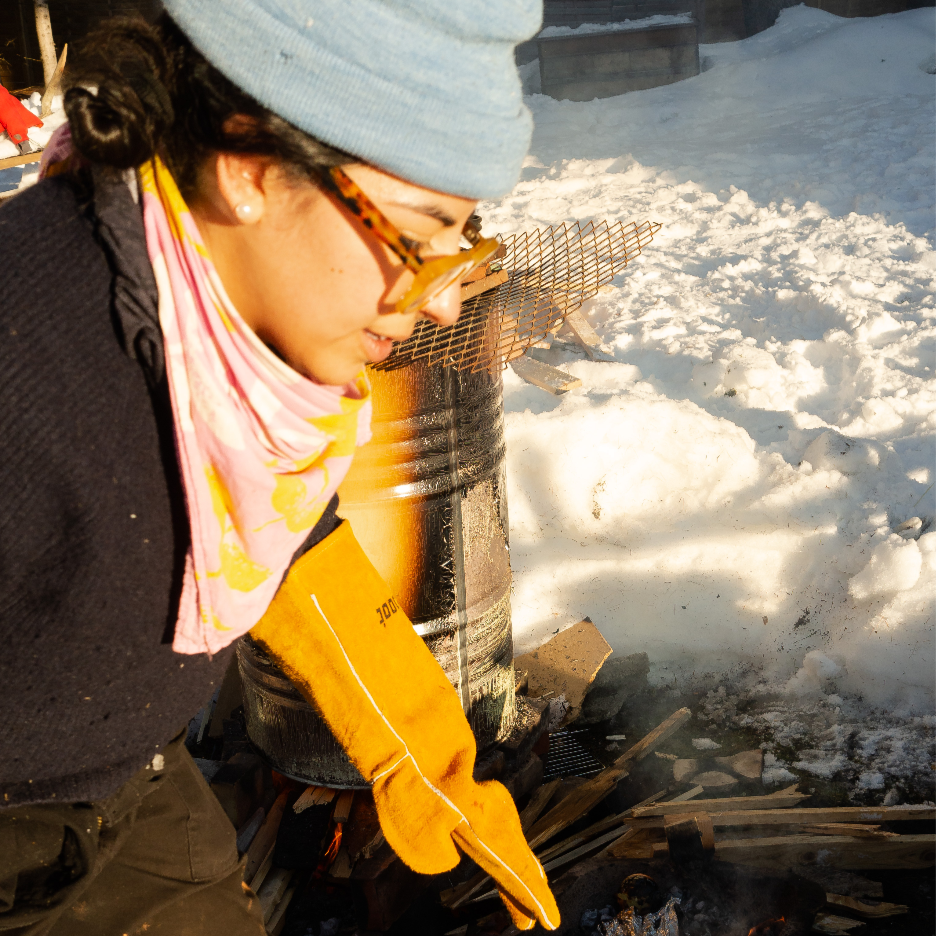 A woman wearing a gray beanie, glasses, and a pink scarf is tending to a fire outdoors on a snowy day, with snow-covered ground and a metal fire pit in the background.