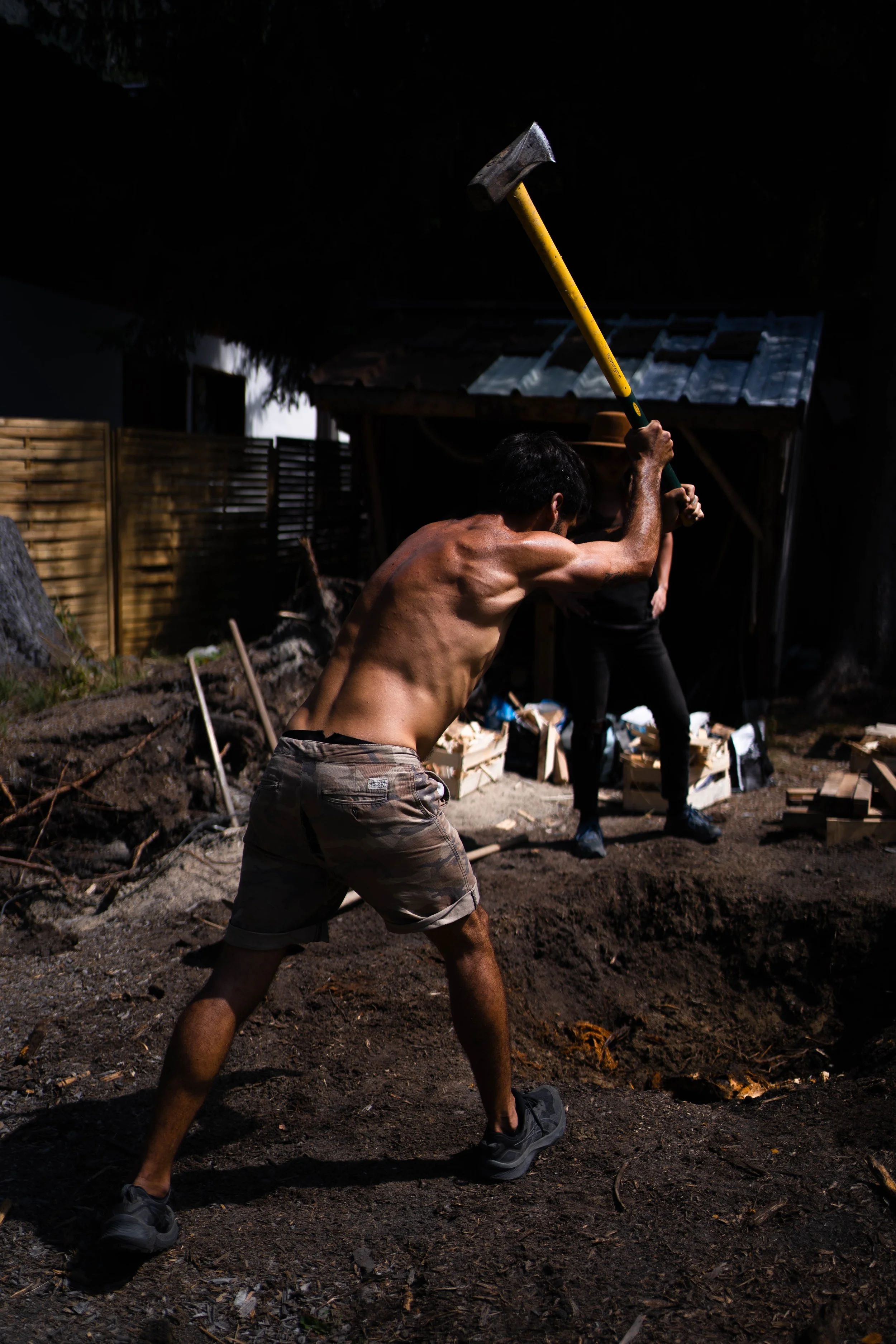 A shirtless man wearing shorts and running shoes is swinging a hammer at the ground in a backyard or construction site at night, with a woman standing in the background and a wooden fence and shed nearby.