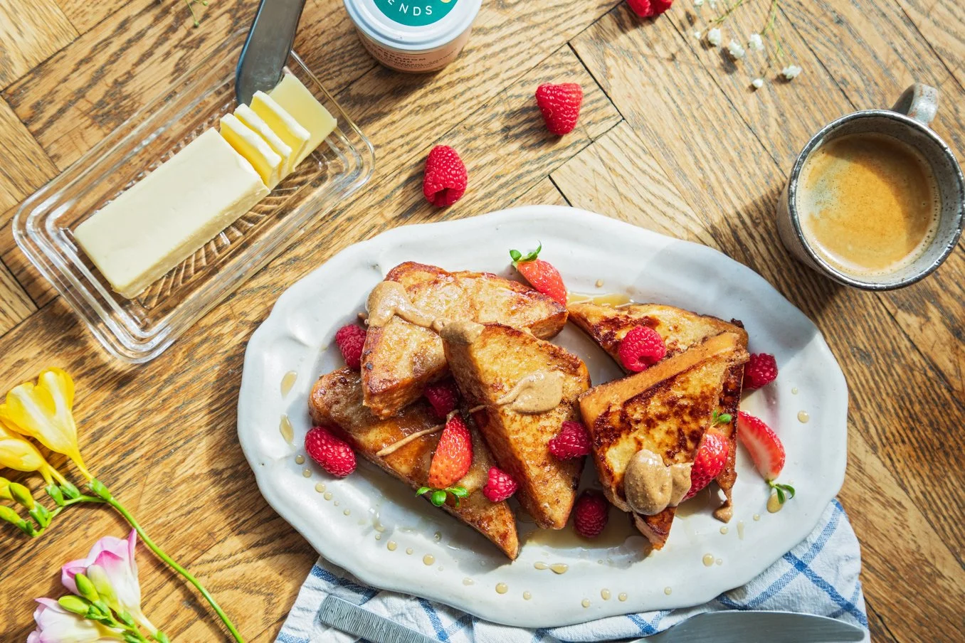 French toast with strawberries and raspberries on a white plate, butter, coffee, and a butter dish on a wooden table