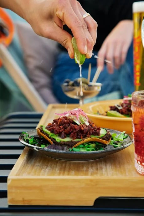 Person squeezing lime over a small plate of tacos with meat, shredded lettuce, and pink onion on a wooden serving board at a restaurant.
