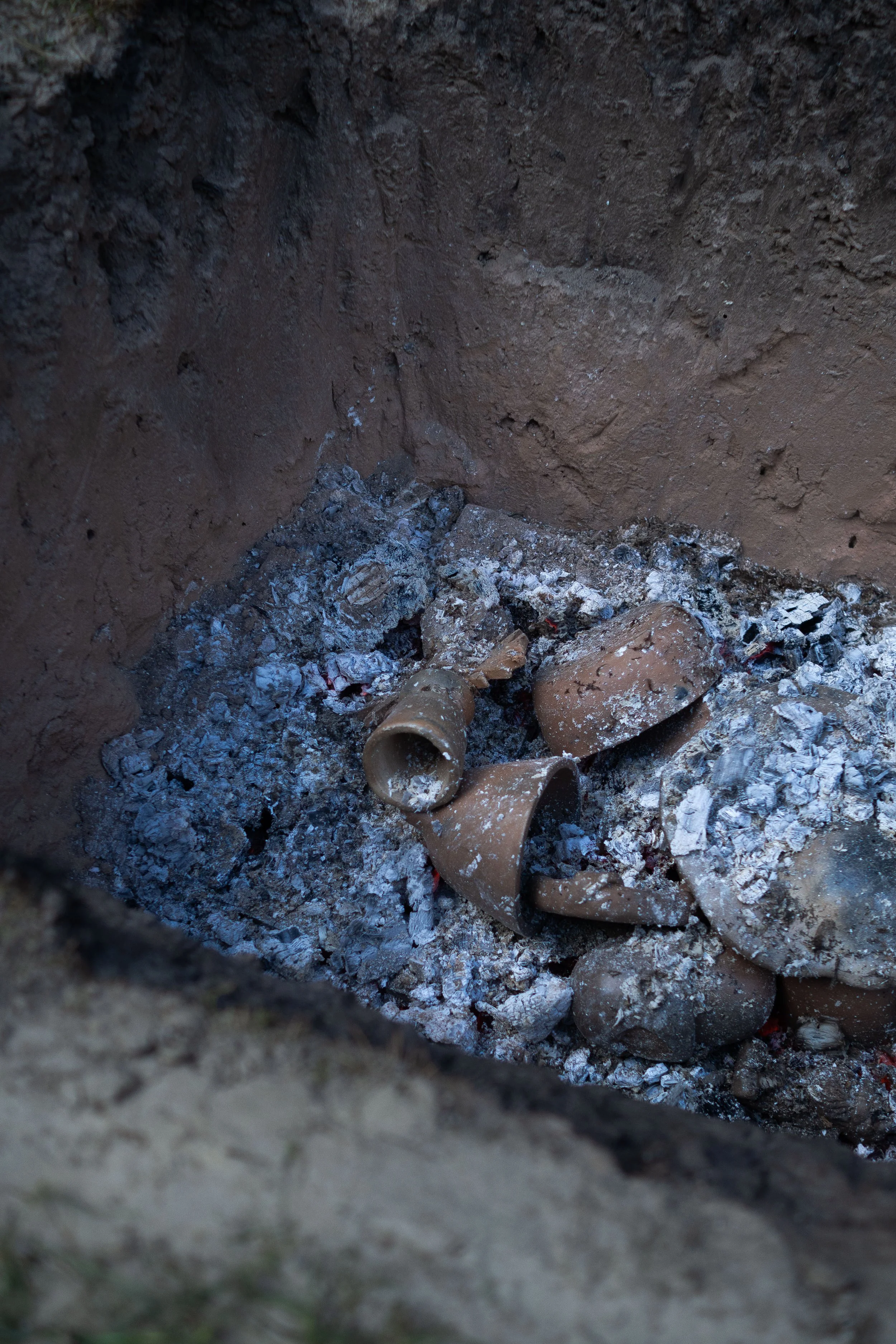 An open fire pit with ash and broken pieces of pottery or ceramic items inside, surrounded by dirt walls.