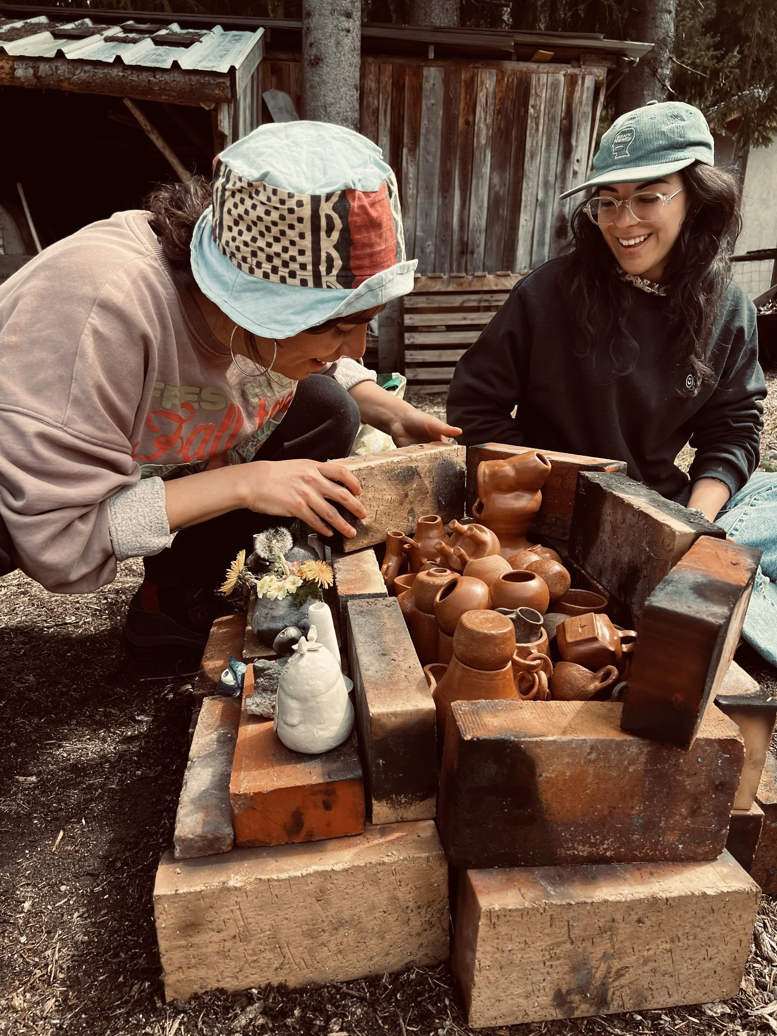 Two women are working on pottery inside a kiln outdoors, surrounded by trees and wooden structures.