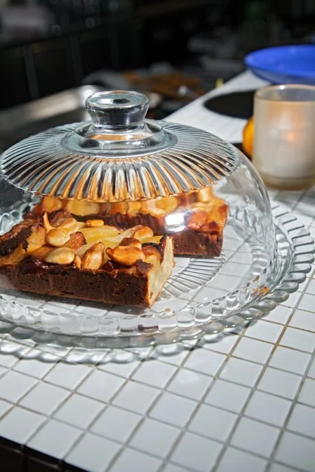 A slice of chocolate brownie with nuts under a glass cake dome on a white tiled surface.