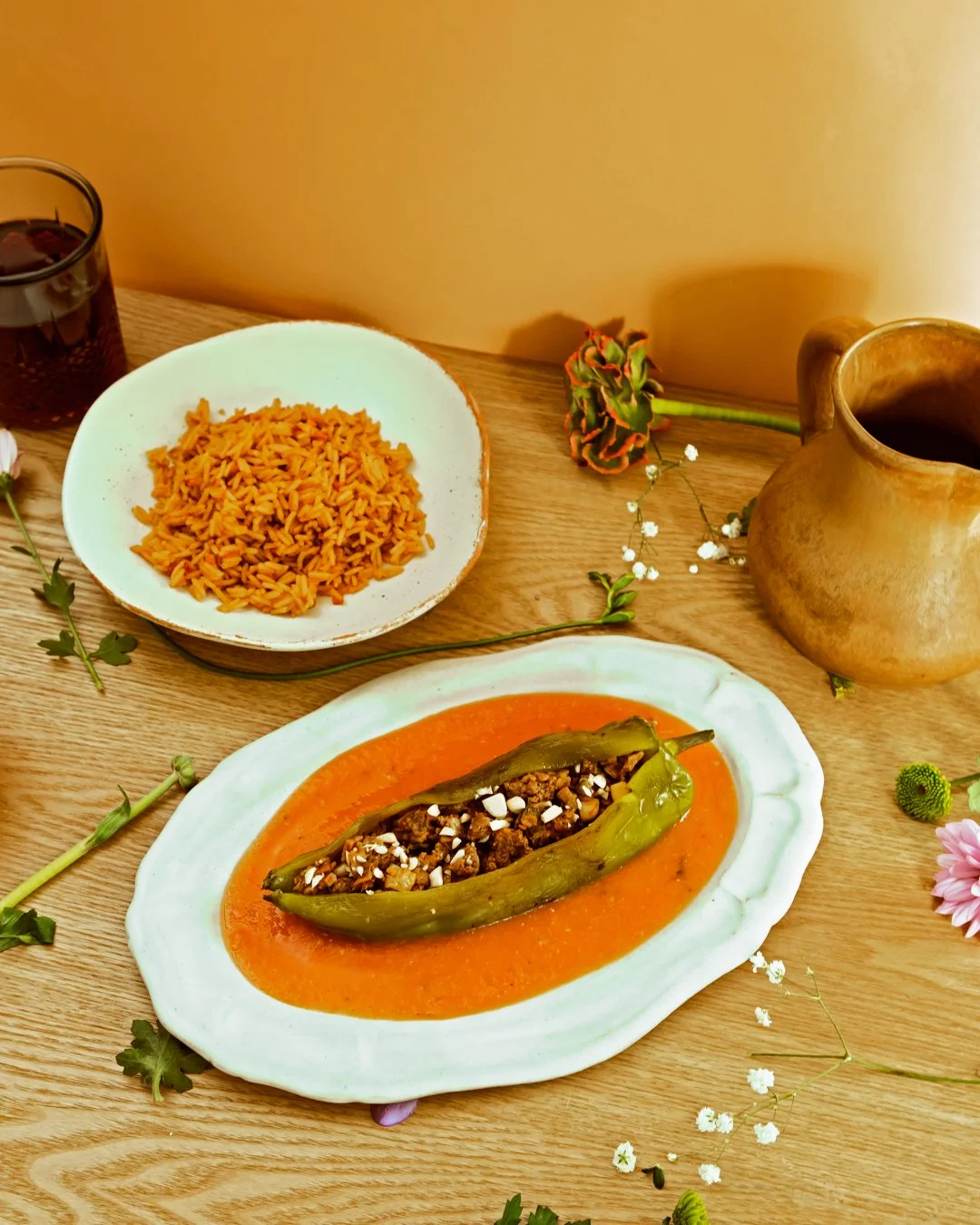 Traditional Mexican meal on a wooden table, featuring a stuffed poblano pepper in red sauce, rice, and a glass of soda, decorated with flowers.