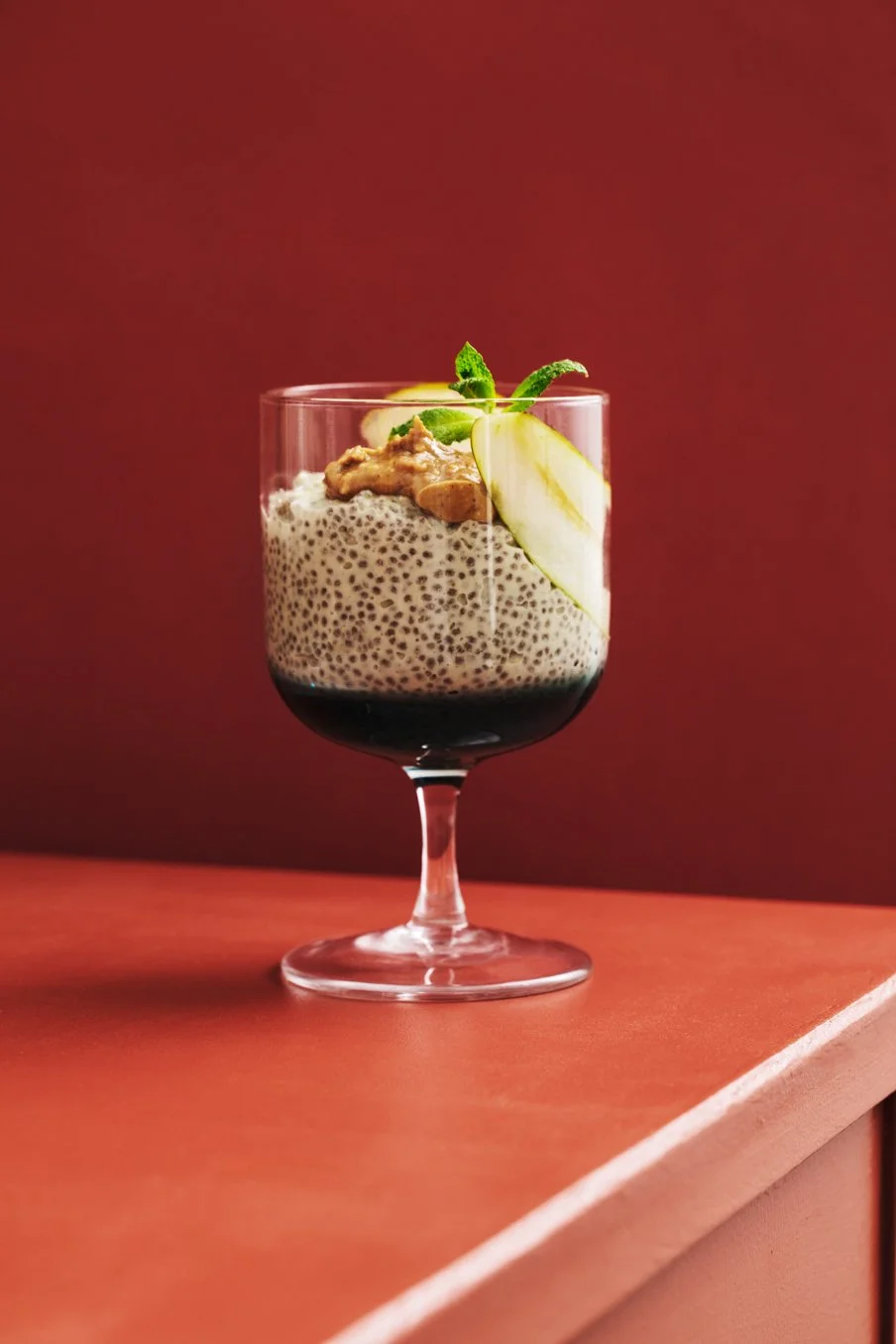 A dessert served in a wine glass with a chia seed pudding base, topped with slices of apple, a dollop of peanut butter or caramel, and garnished with a mint leaf, set against a reddish-brown background.