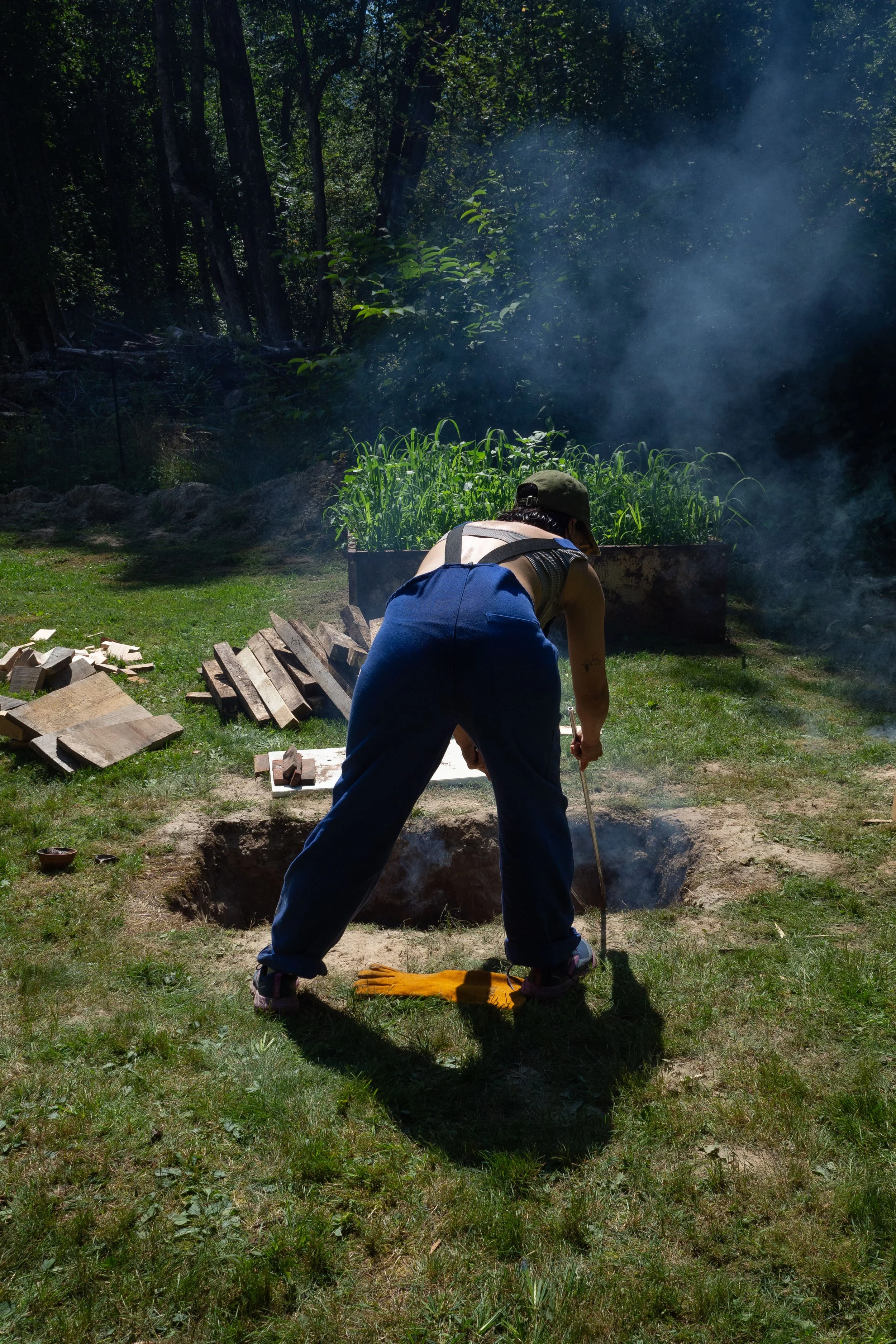 Person wearing a cap and sleeveless shirt working in a garden, pushing soil with a tool, with smoke rising and a garden bed with plants in the background.