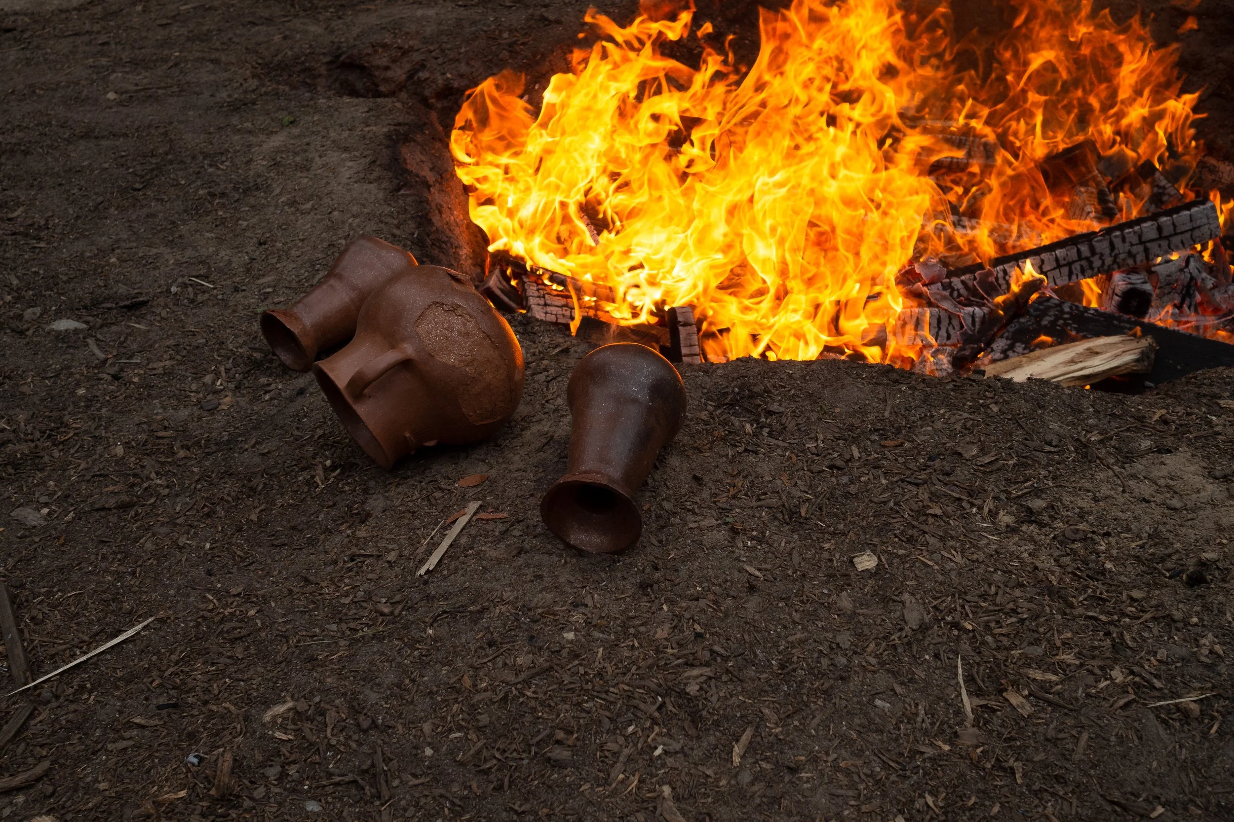 A large fire burns on the ground with bright orange flames and burning wood. Several broken pottery pieces lie on the dirt near the fire.