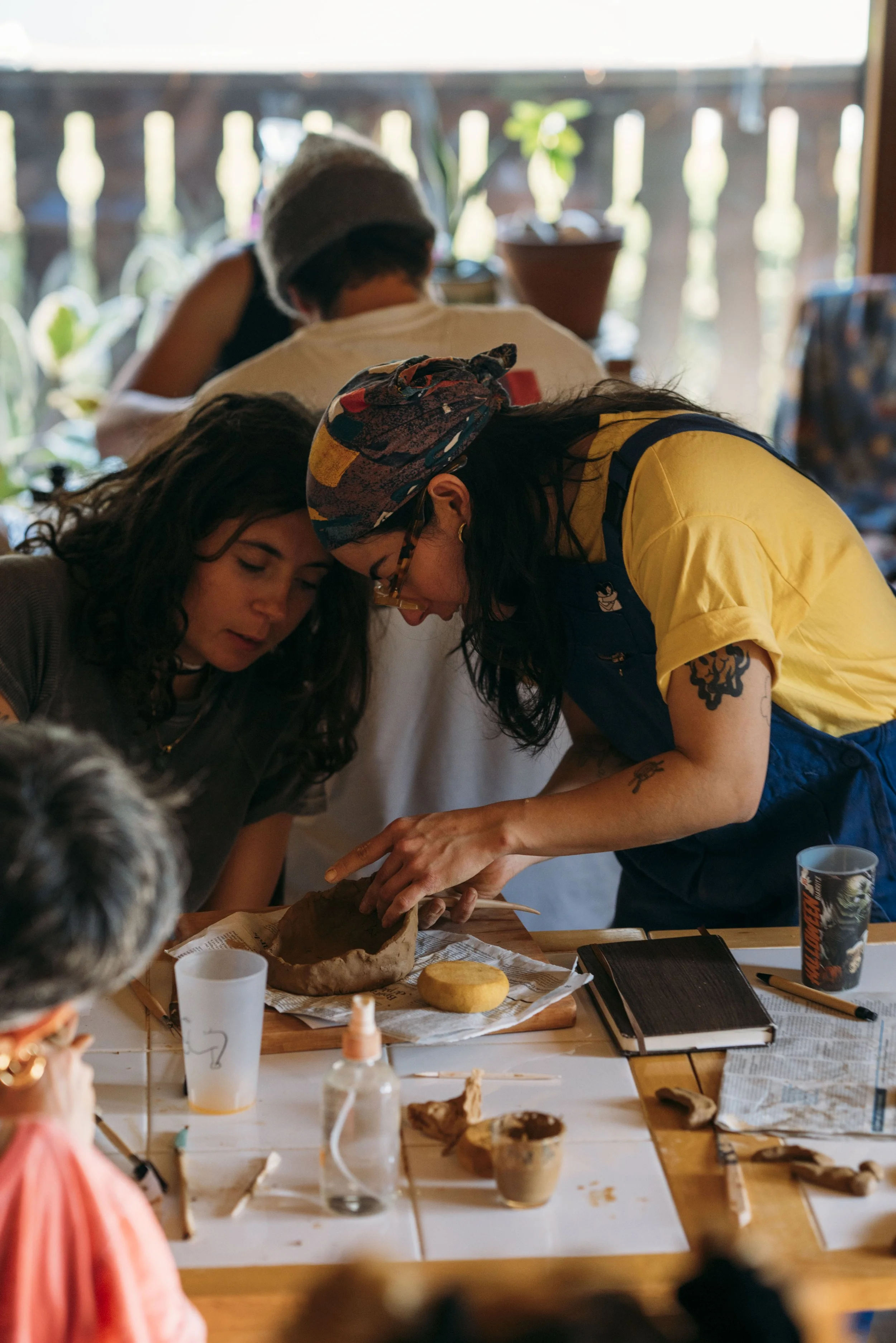 Two women with long dark hair working together on a project with clay, at a table with tools, books, and drinks, in a room with a window and plants in the background.