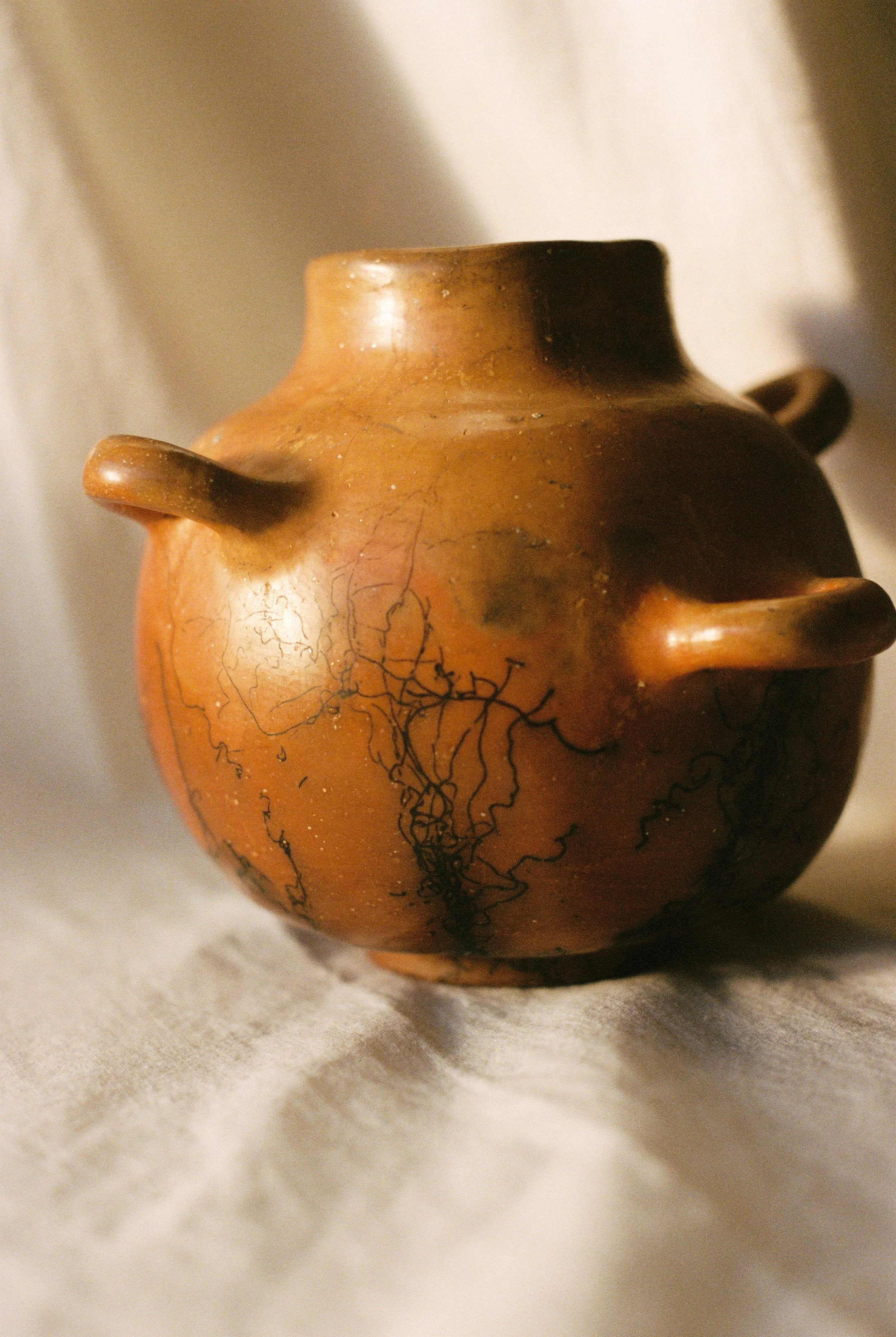 Close-up of an old, round, brown ceramic pot with three small handles, resting on a soft, light-colored fabric surface.