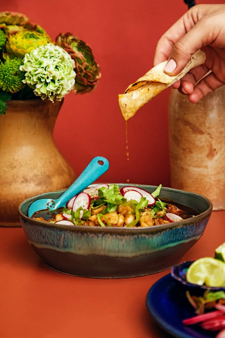 Person dipping a tortilla chip into a bowl of soup or chili garnished with sliced radishes and green onions, with a vase of succulents and a plate of lime slices in the background.