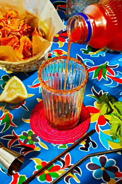 Pouring a red soda into a glass on a colorful table with chips, lemon wedge, and herbs.