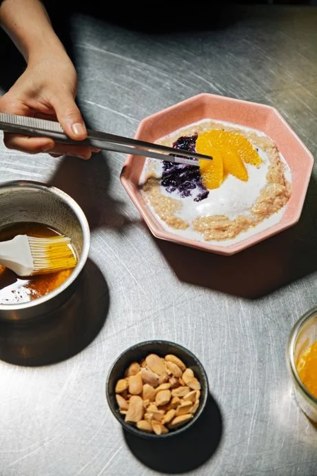 A bowl of oatmeal topped with orange slices, blueberries, and yogurt. A person is using tongs to add more orange slices to the oatmeal. There are small bowls of chopped nuts, honey, and another ingredient nearby.