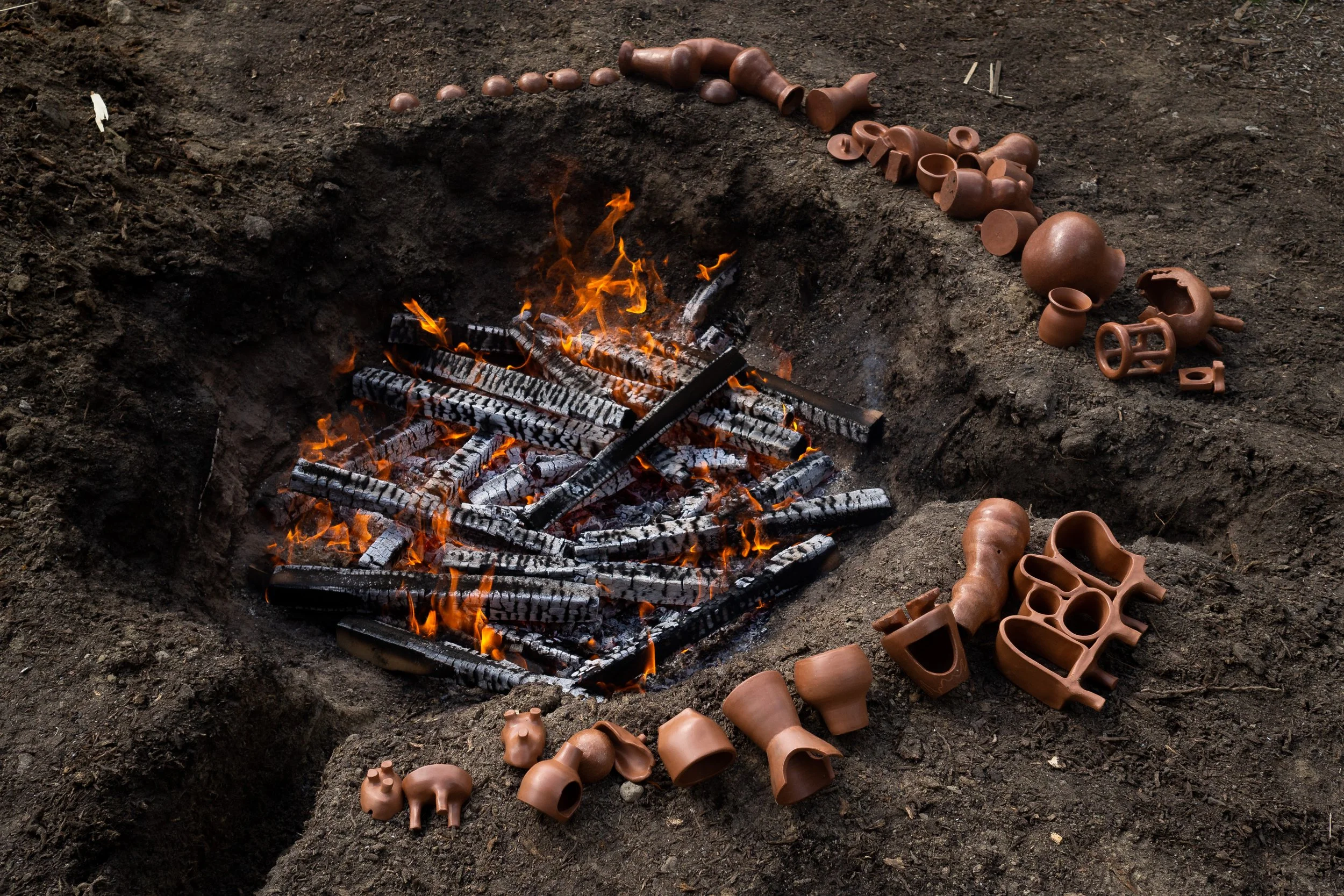 Archaeological excavation site with a pit containing burning charcoal and a variety of ancient clay pottery pieces scattered around the edges.