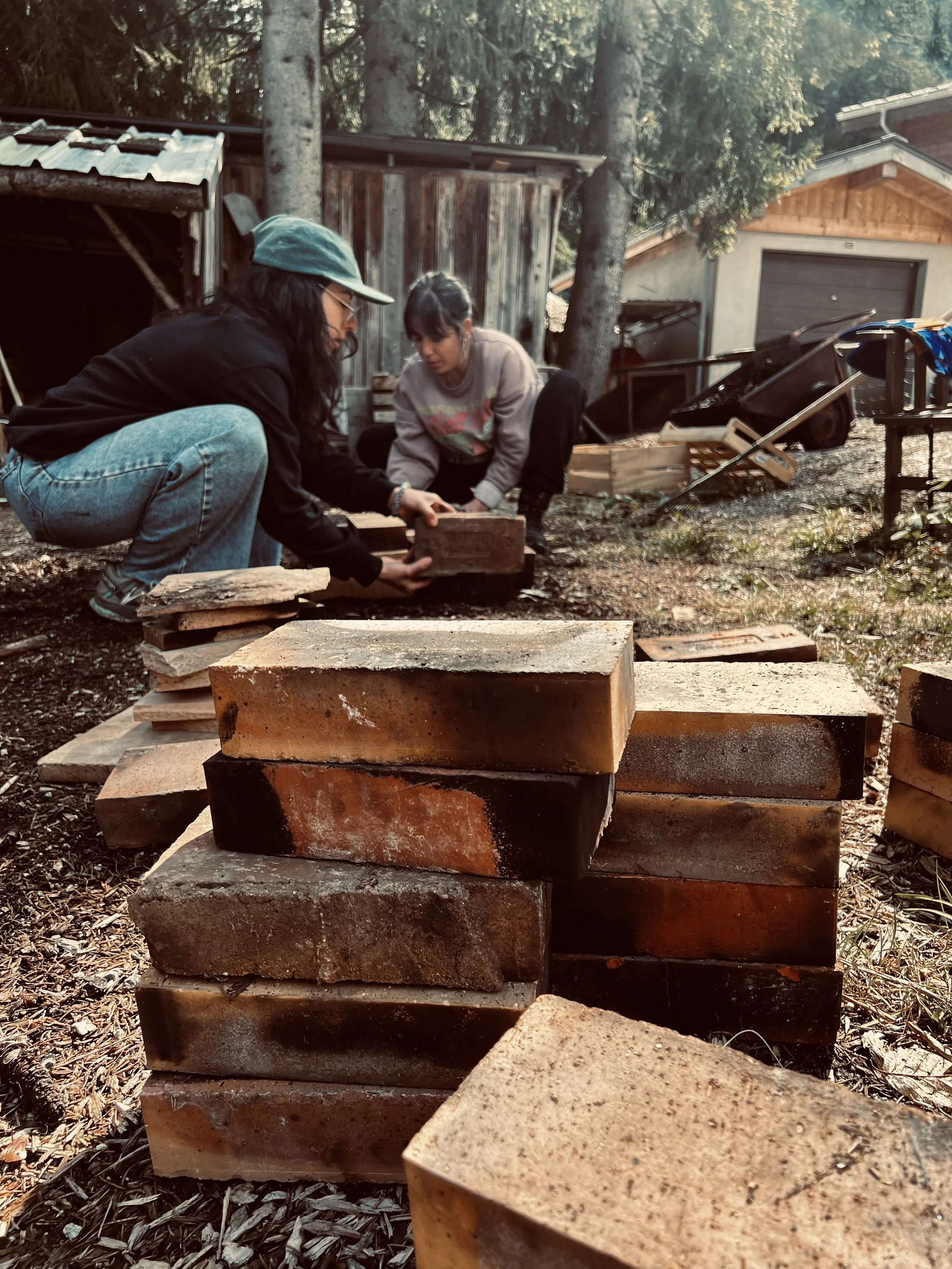 Two women working outdoors in a wooded area with stacked bricks, a wooden shed, and trees in the background.