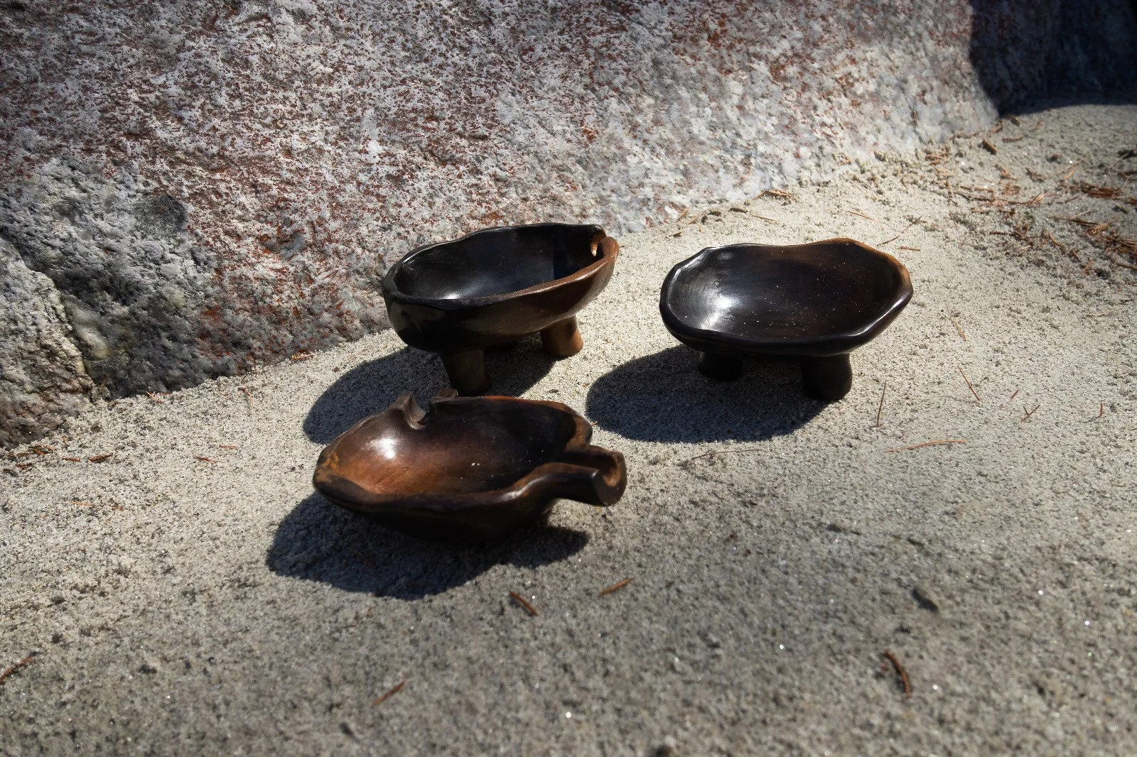 Three small dark wooden bowls on sandy ground near rocks, shadowed in sunlight.