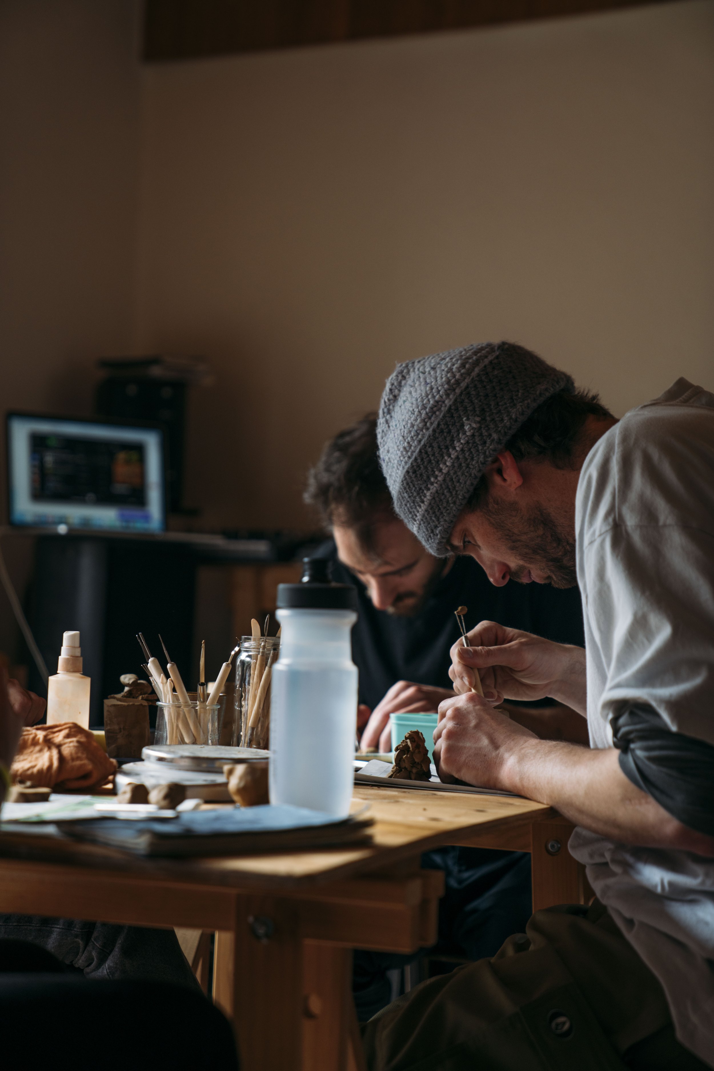 Two men working on a project at a wooden table in a dimly lit room, with various pottery tools and bottles on the table, and a computer in the background.