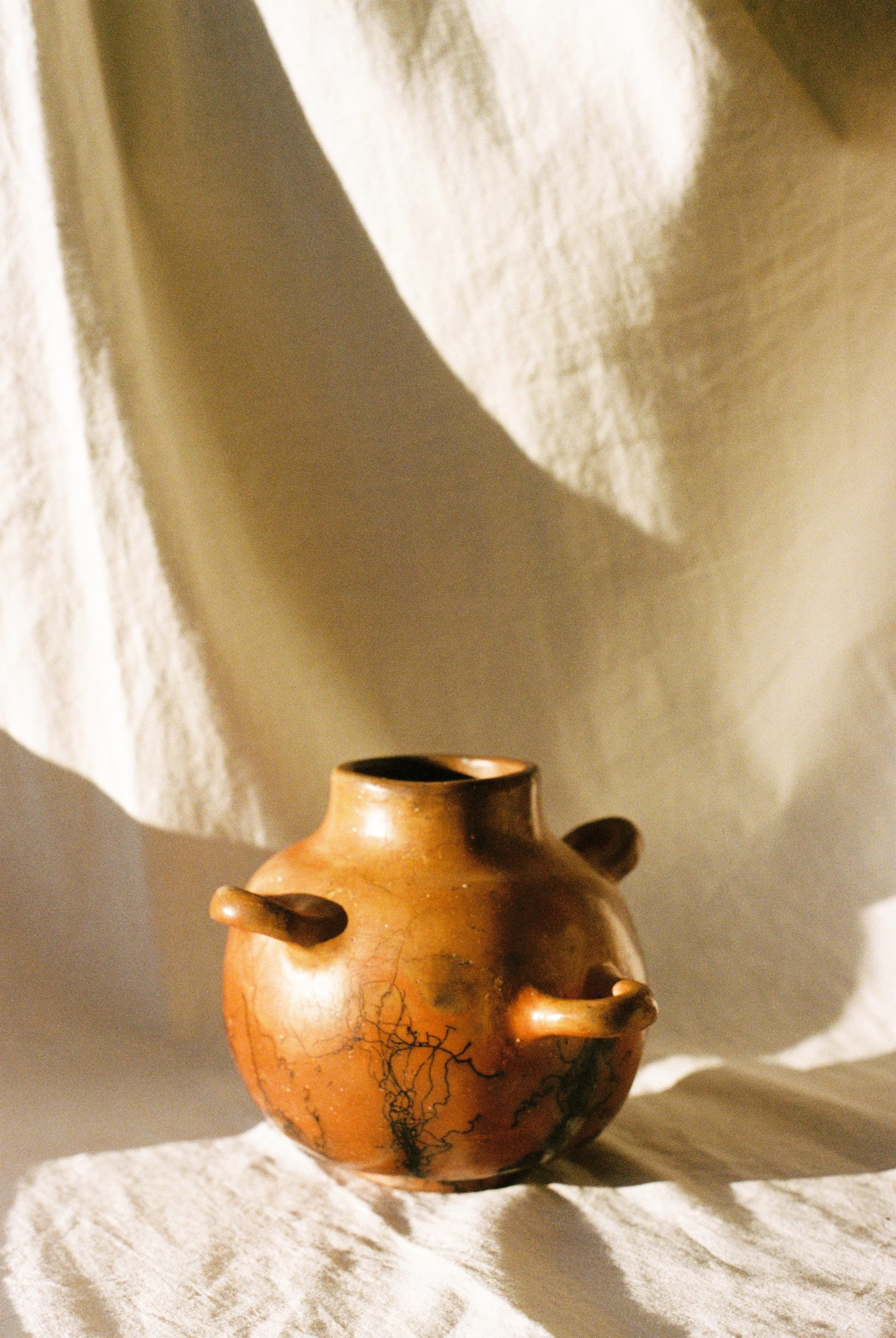 A small, round, terracotta vase with black crackle patterns and decorative protruding arms. The vase is placed on a textured, light-colored fabric backdrop, with a piece of light-colored cloth draped behind it, creating soft shadows.