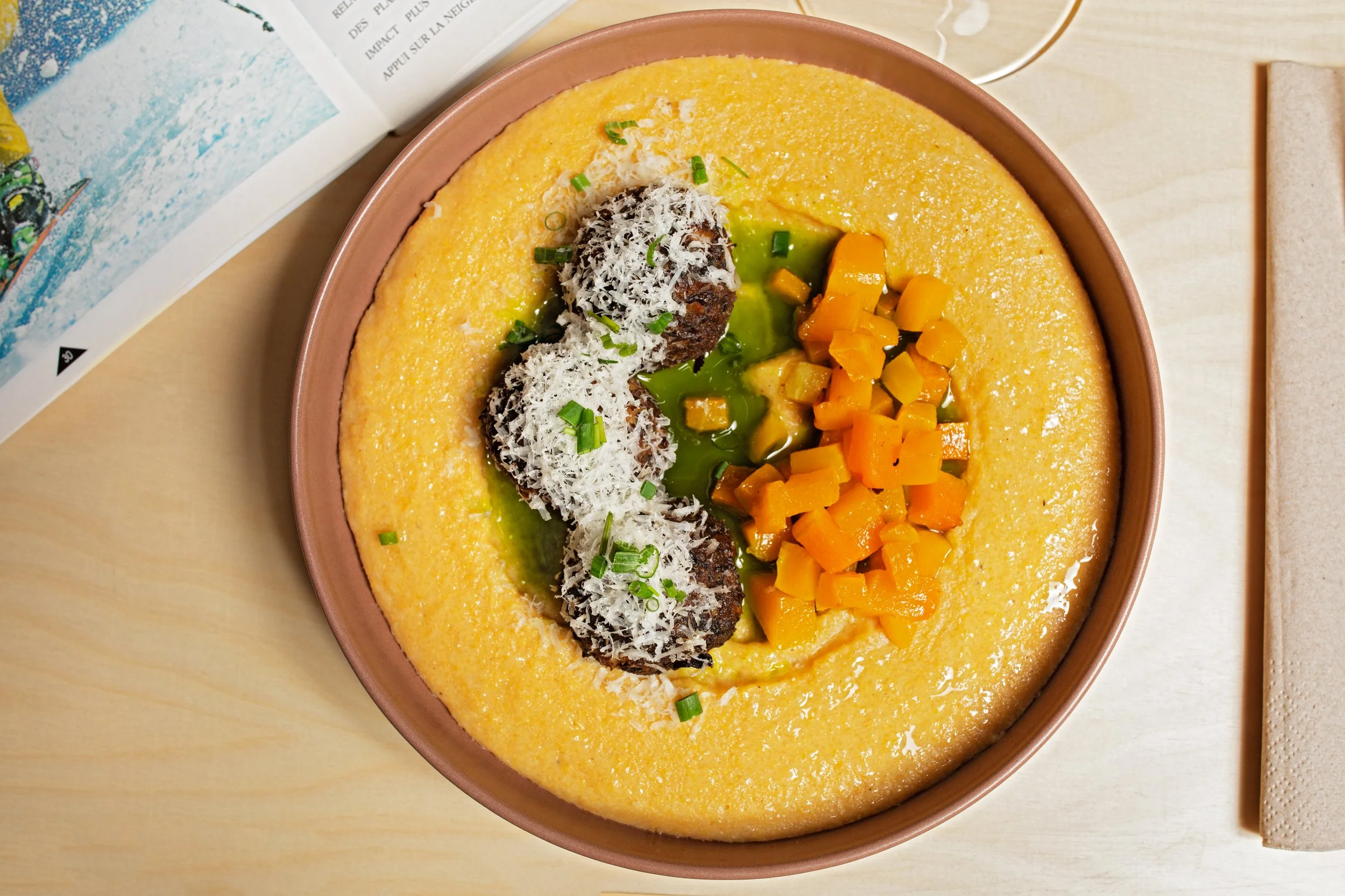 A bowl of creamy yellow corn soup garnished with shredded cheese, chopped green onions, diced carrots, and two meatballs.