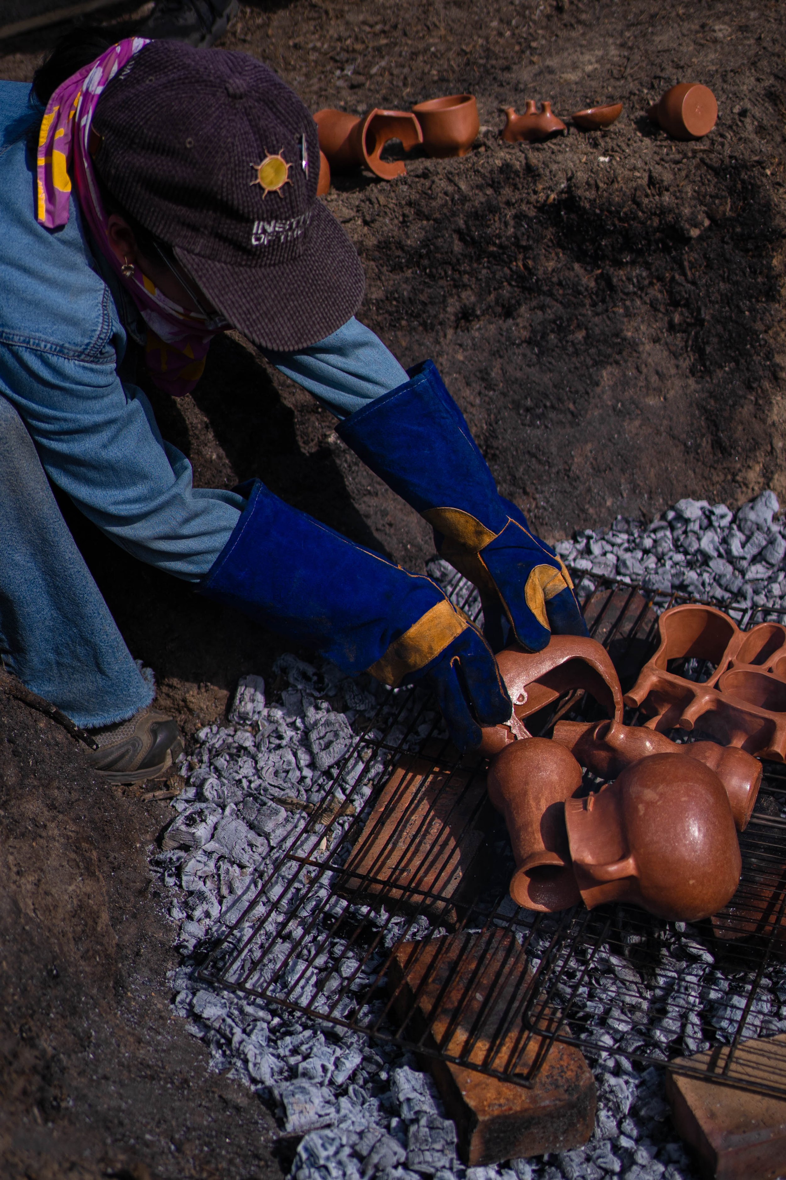 A person working with archaeological artifacts, including pottery shards and figurines, on a wire rack over a bed of ashes.