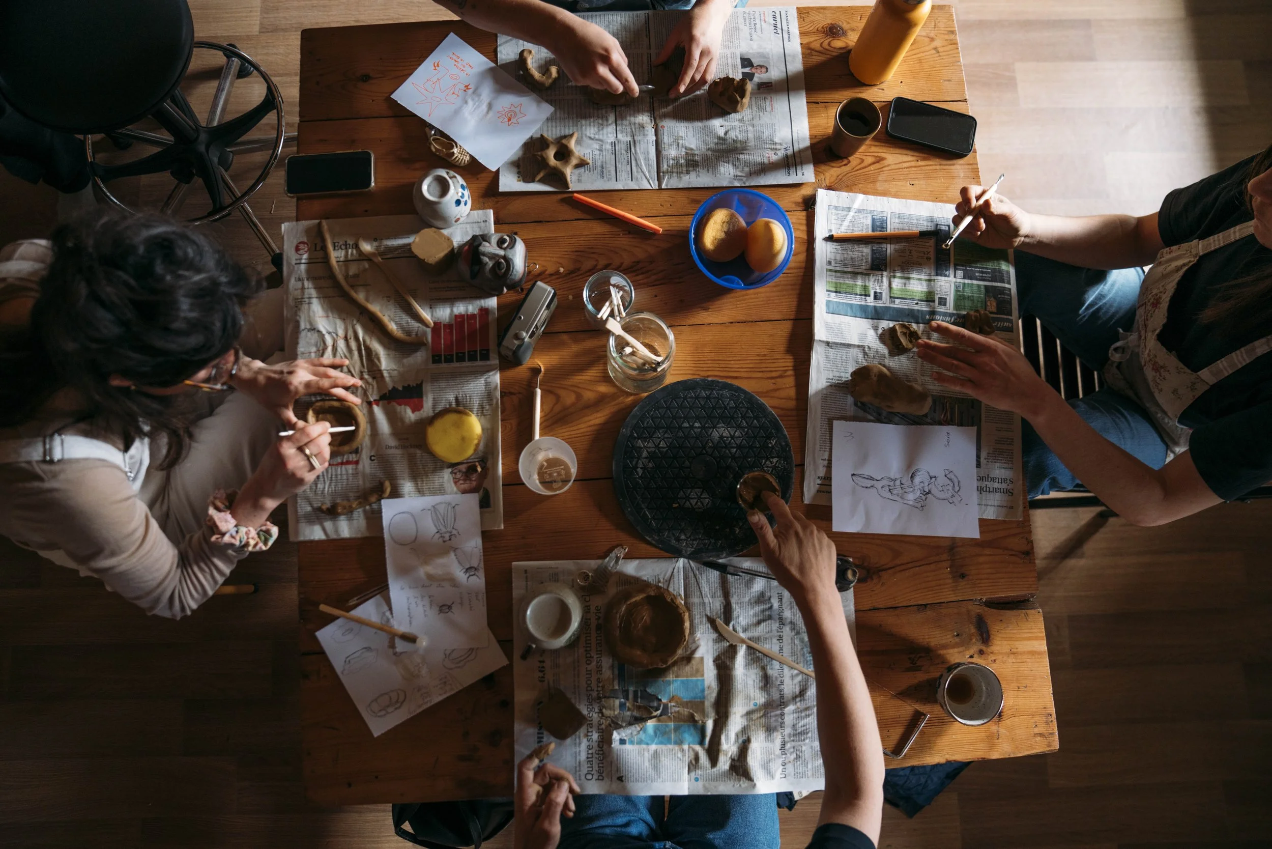 A top-down view of a wooden table covered with newspapers, paper sketches, clay sculptures, and art supplies. Several people are working on craft projects, shaping clay and drawing. Cups of coffee and a bowl with yellow sponges are also visible.