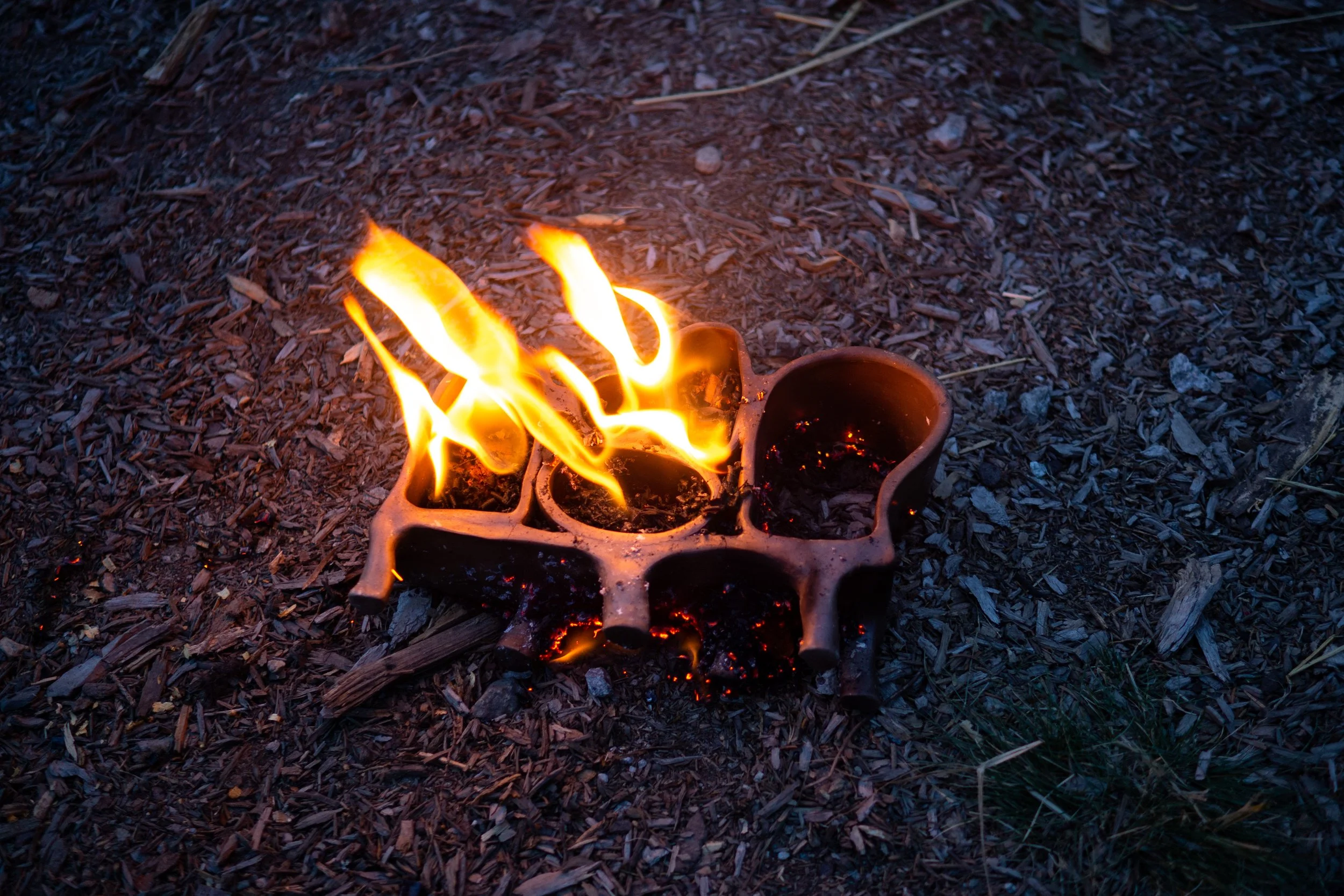 A ceramic mold with compartments filled with hot, glowing coals, placed on a bed of wood chips, with flames emerging from the coals.