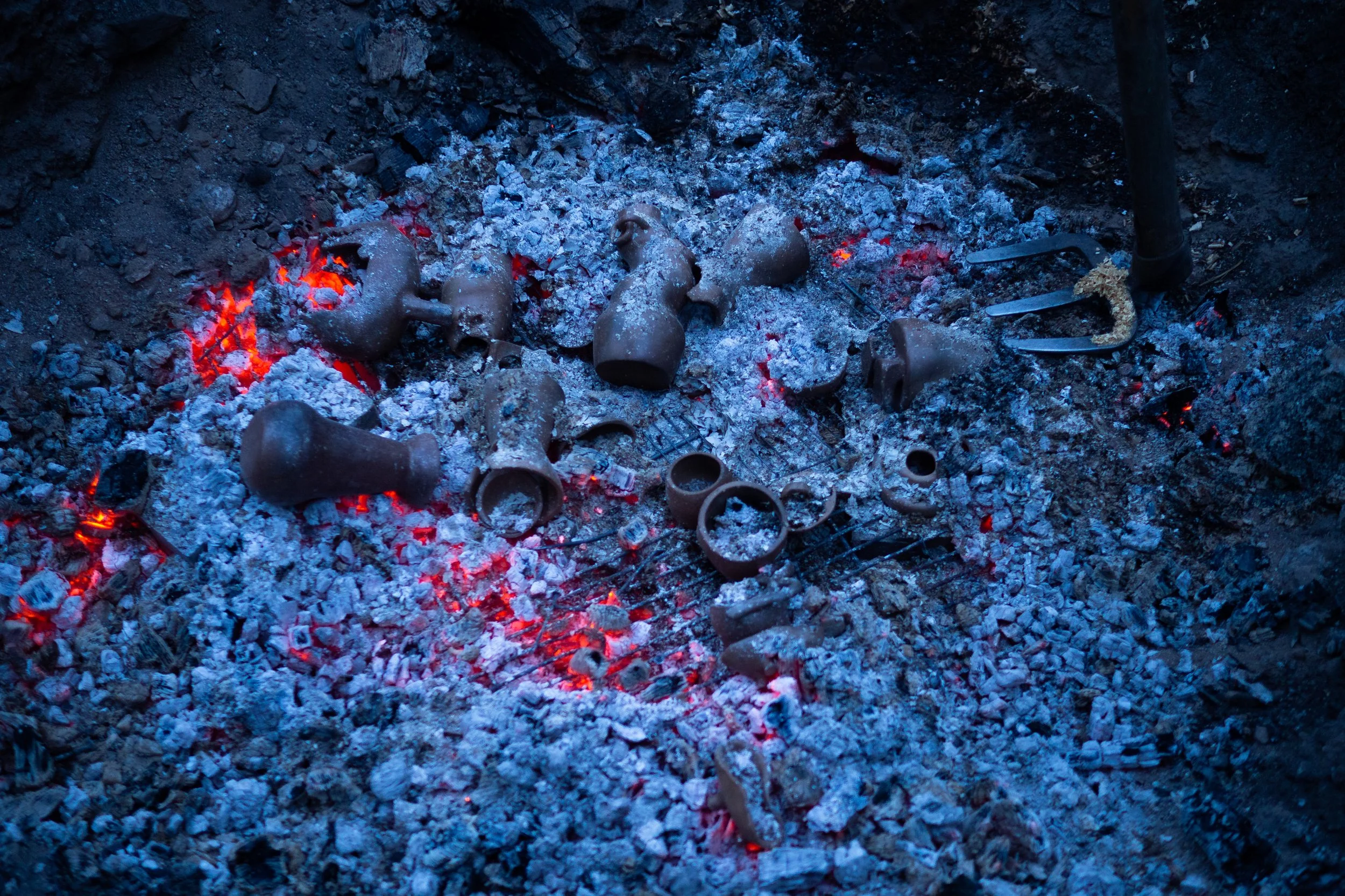 Hot, glowing embers and ash from a fire, with scattered ceramic pieces and metal tools.
