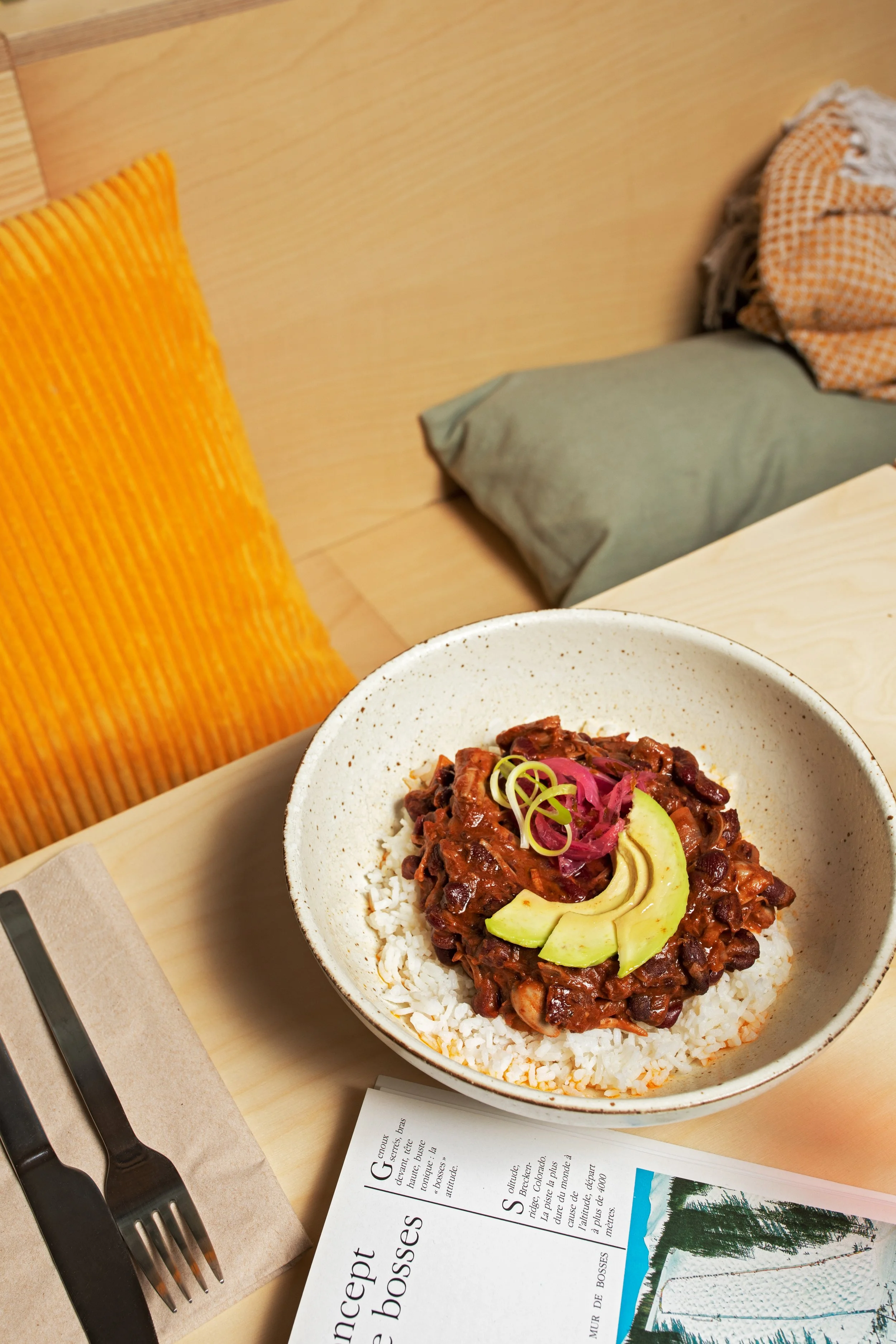 A bowl of rice topped with a dark red meat and vegetable stew, garnished with sliced avocado and pickled red onions, on a wooden tray with a black fork and a white paper napkin, next to a newspaper and a spoon. In the background, there are a yellow p
